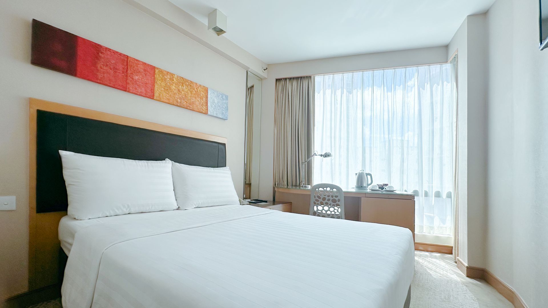 Hotel bedroom with a bed, desk, and a large window overlooking a city.