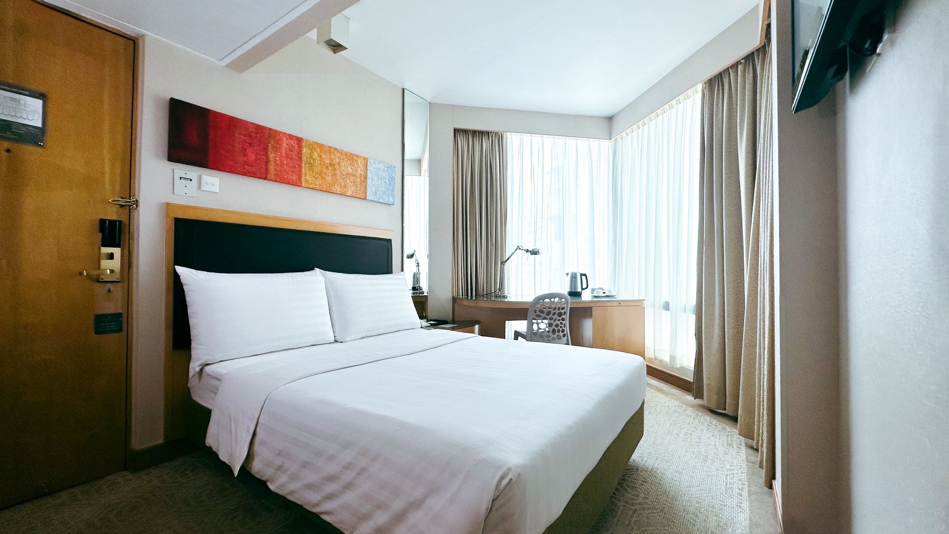 Hotel room with bed, desk by window, curtains, artwork, and TV on wall.
