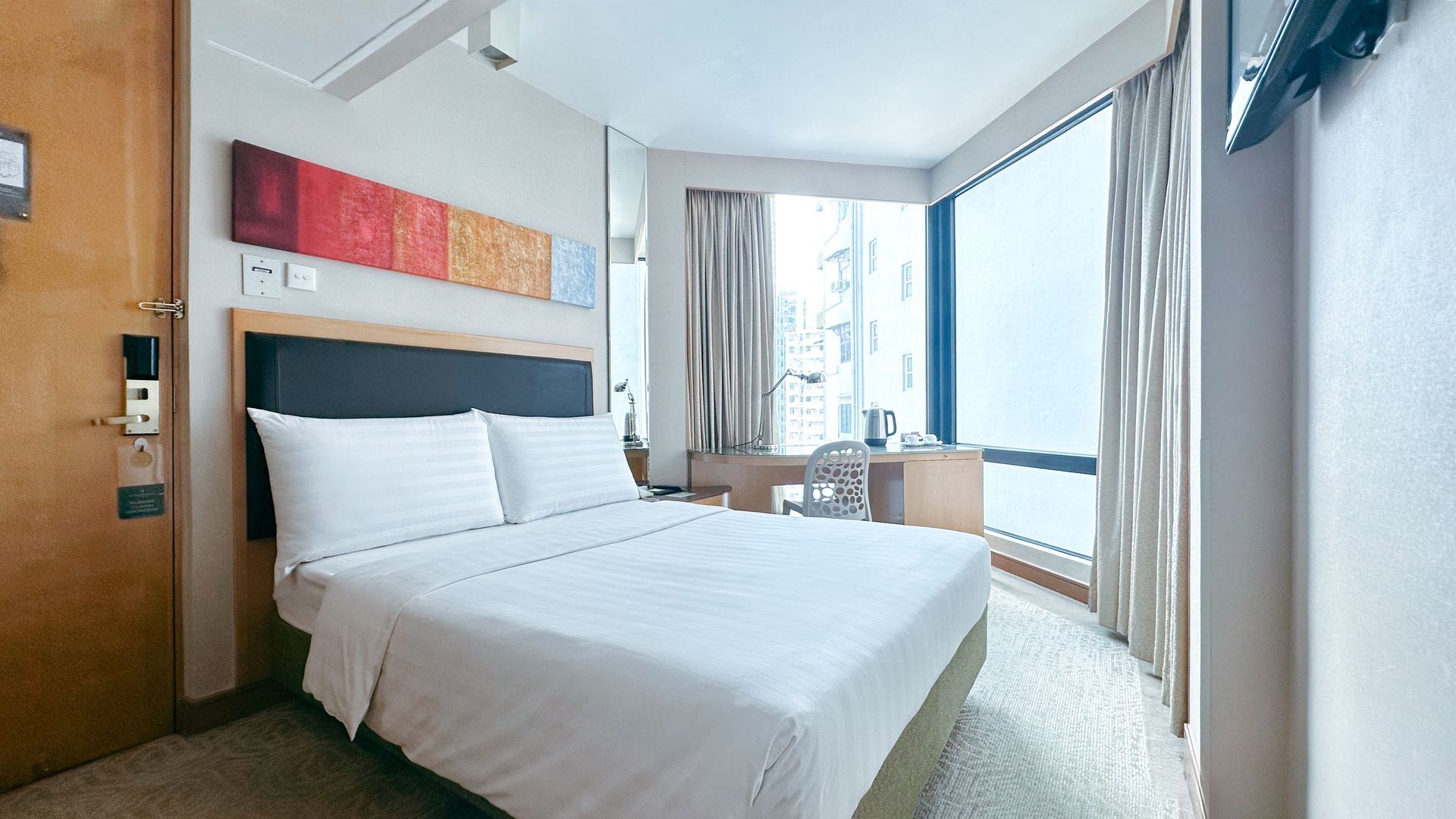 Hotel bedroom with a bed, desk, and large window with a city view.
