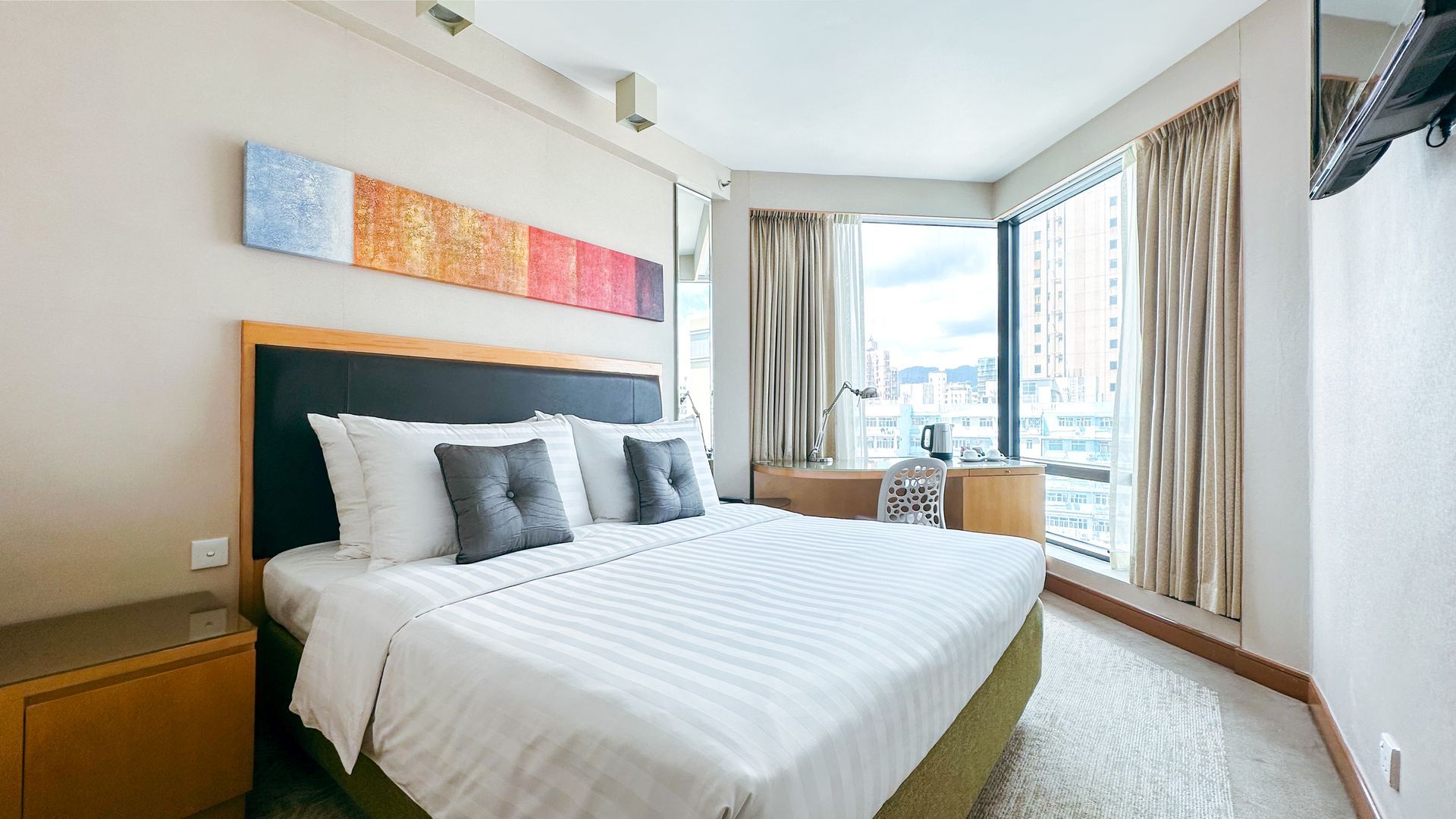 明亮的臥室，床鋪整齊，牆上裝飾著色彩鮮豔的藝術品，透過大窗戶可以欣賞到城市景觀。