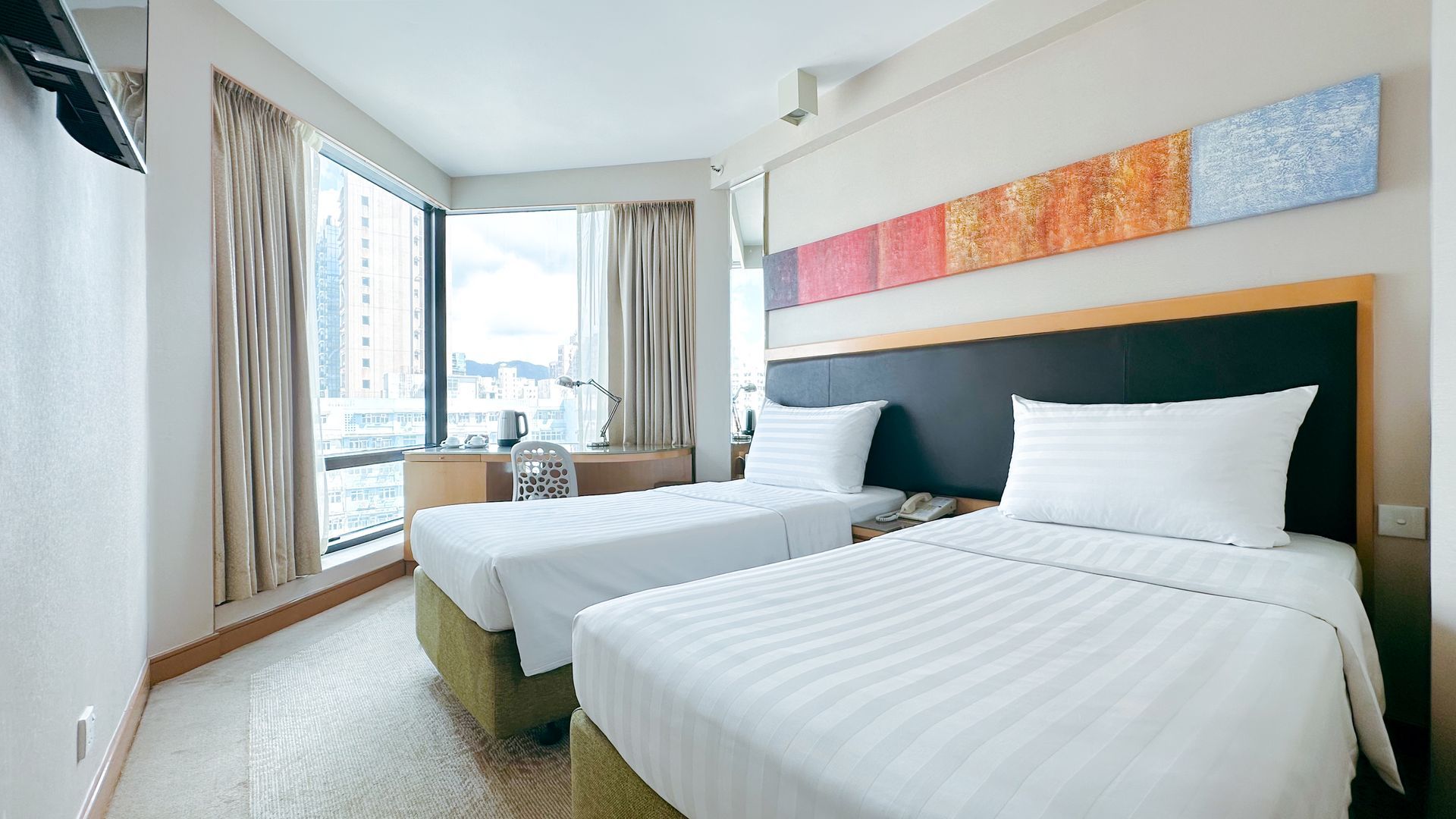 Twin beds in a hotel room with a desk, window, and floral painting.