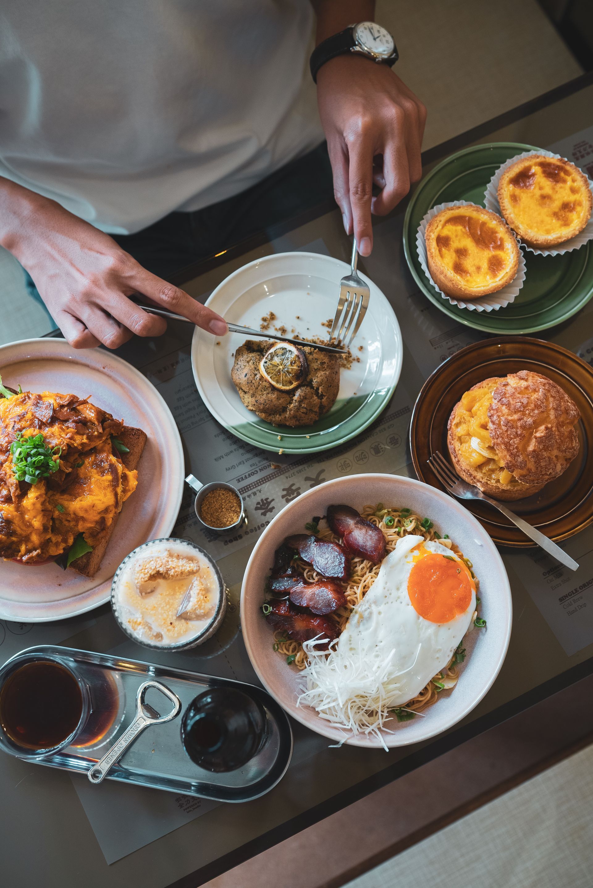 Person cutting into a pastry surrounded by dishes like egg tarts, fried rice, and coffee on a table.