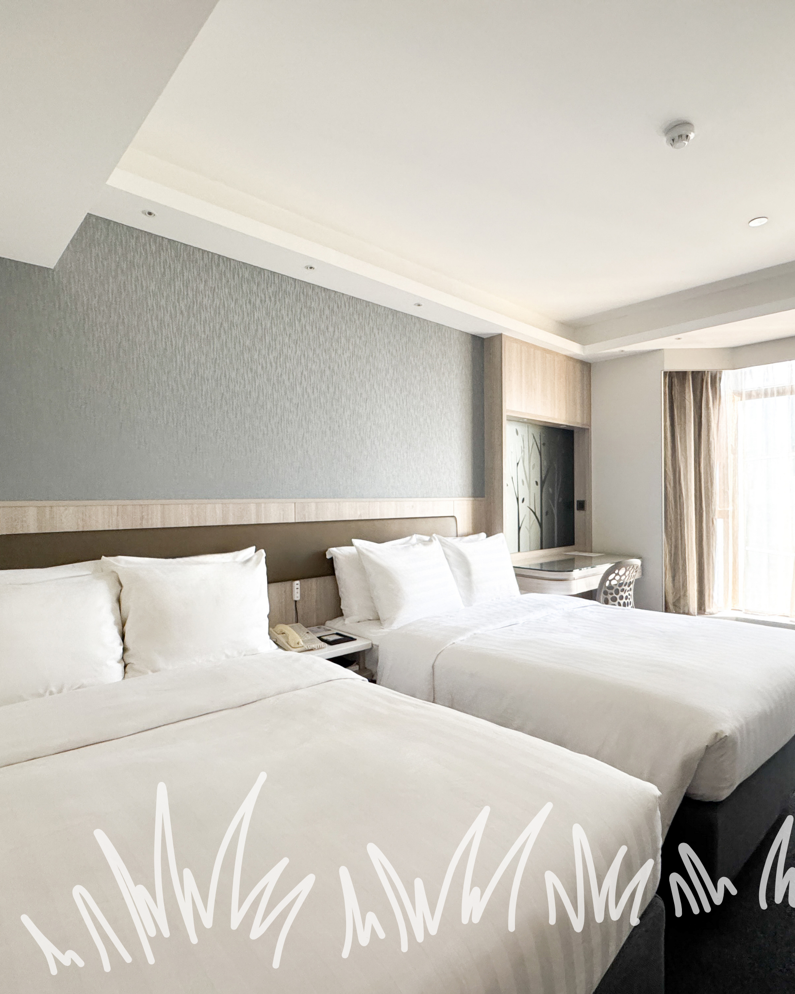 A hotel room with two white beds and a shared nightstand, decorated with white chalk-style drawings of hanging eggs.