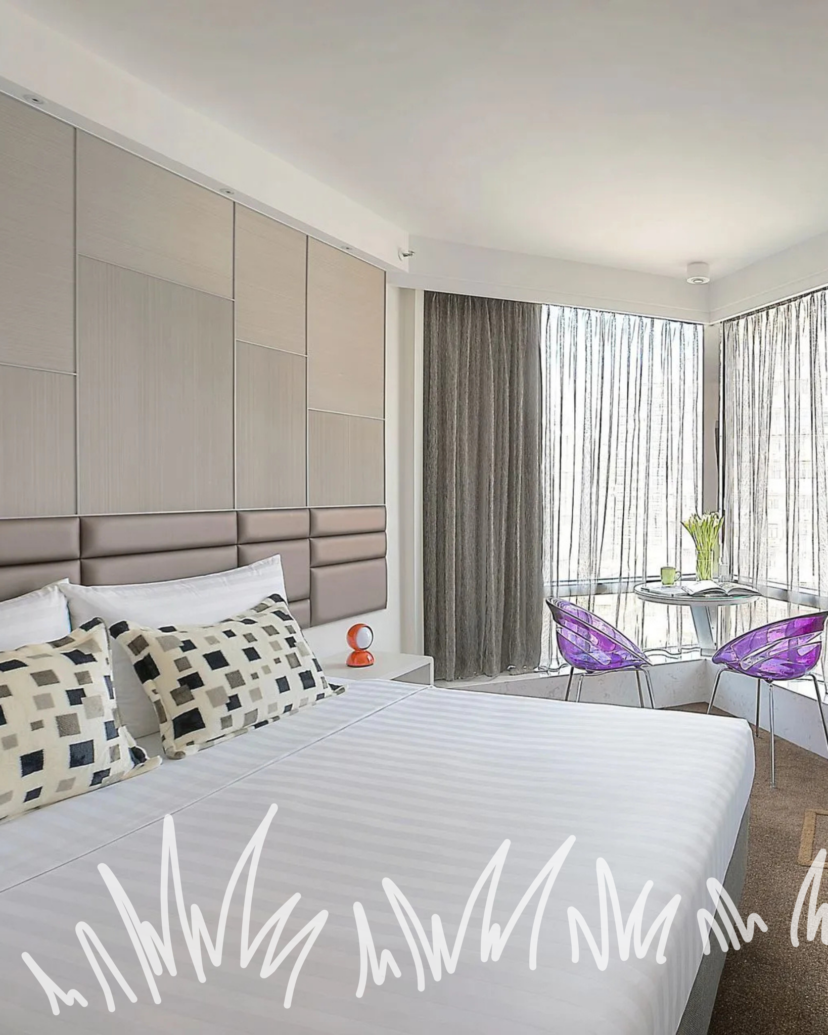 A bedroom features a white-covered bed with patterned pillows against a segmented wall, adjacent to a window with drapes.