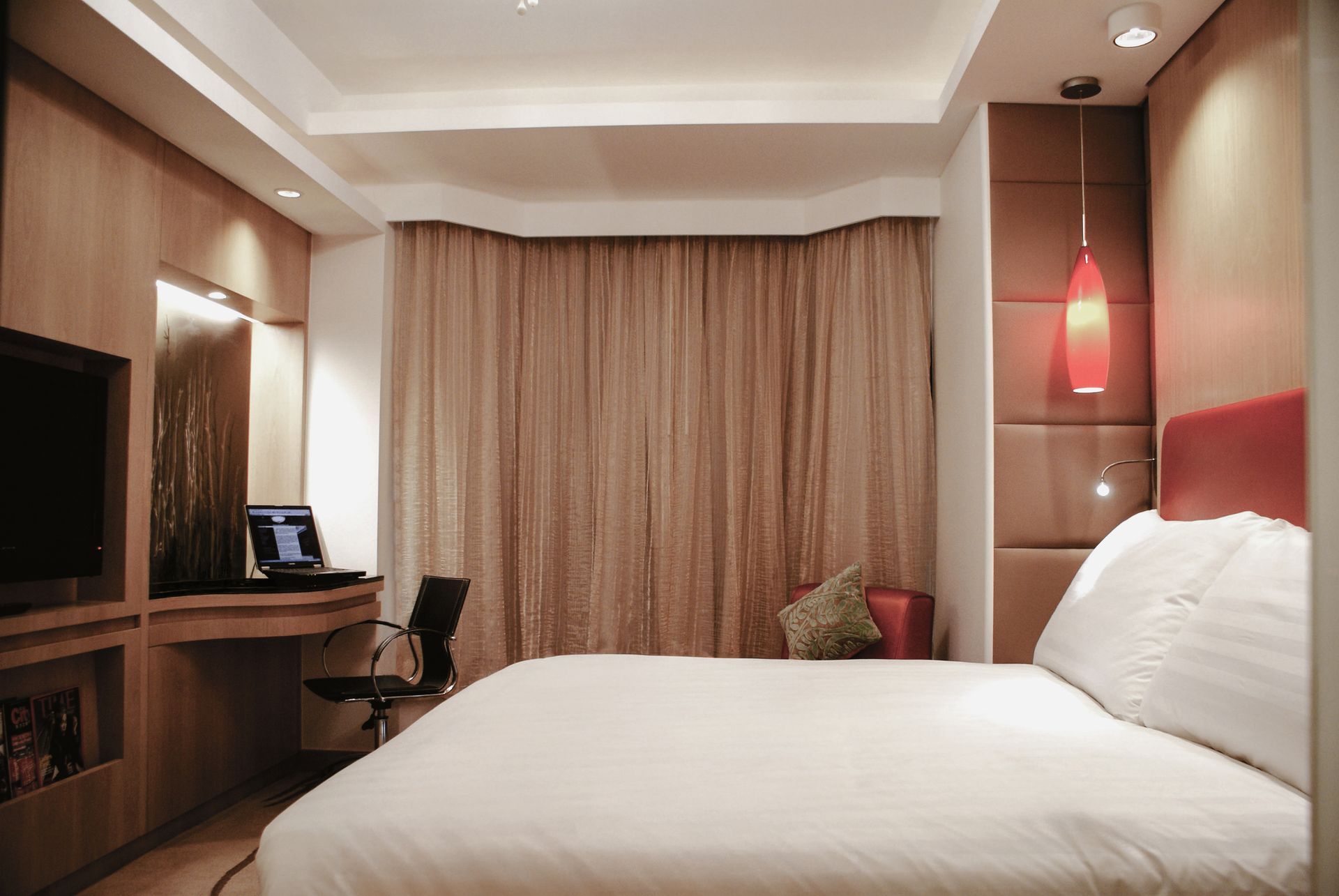 Hotel room with a bed, desk, and TV. Beige and brown tones, with a red headboard and accent light.