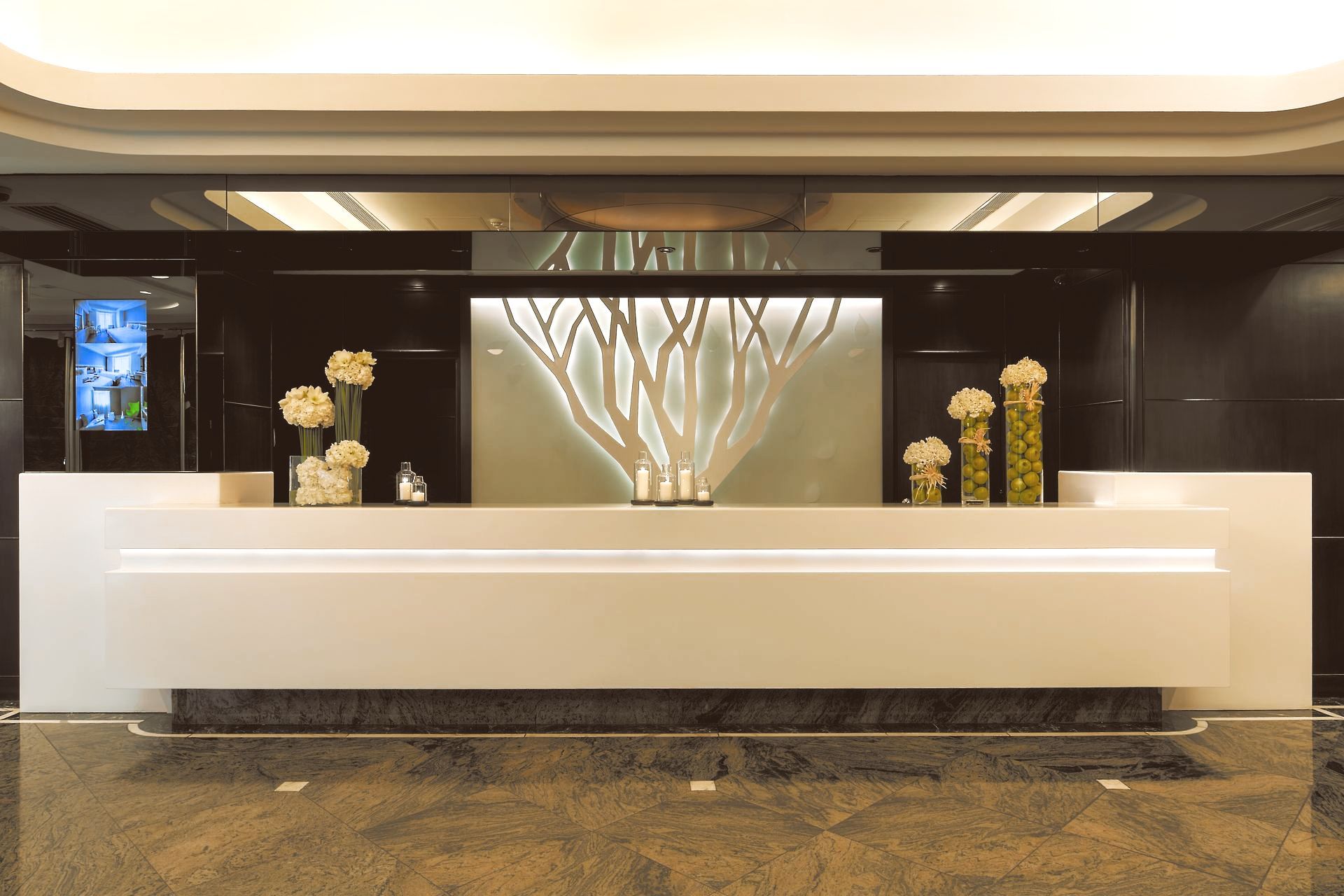 White reception desk with floral arrangements, a decorative light fixture, and a marble floor in a hotel lobby.