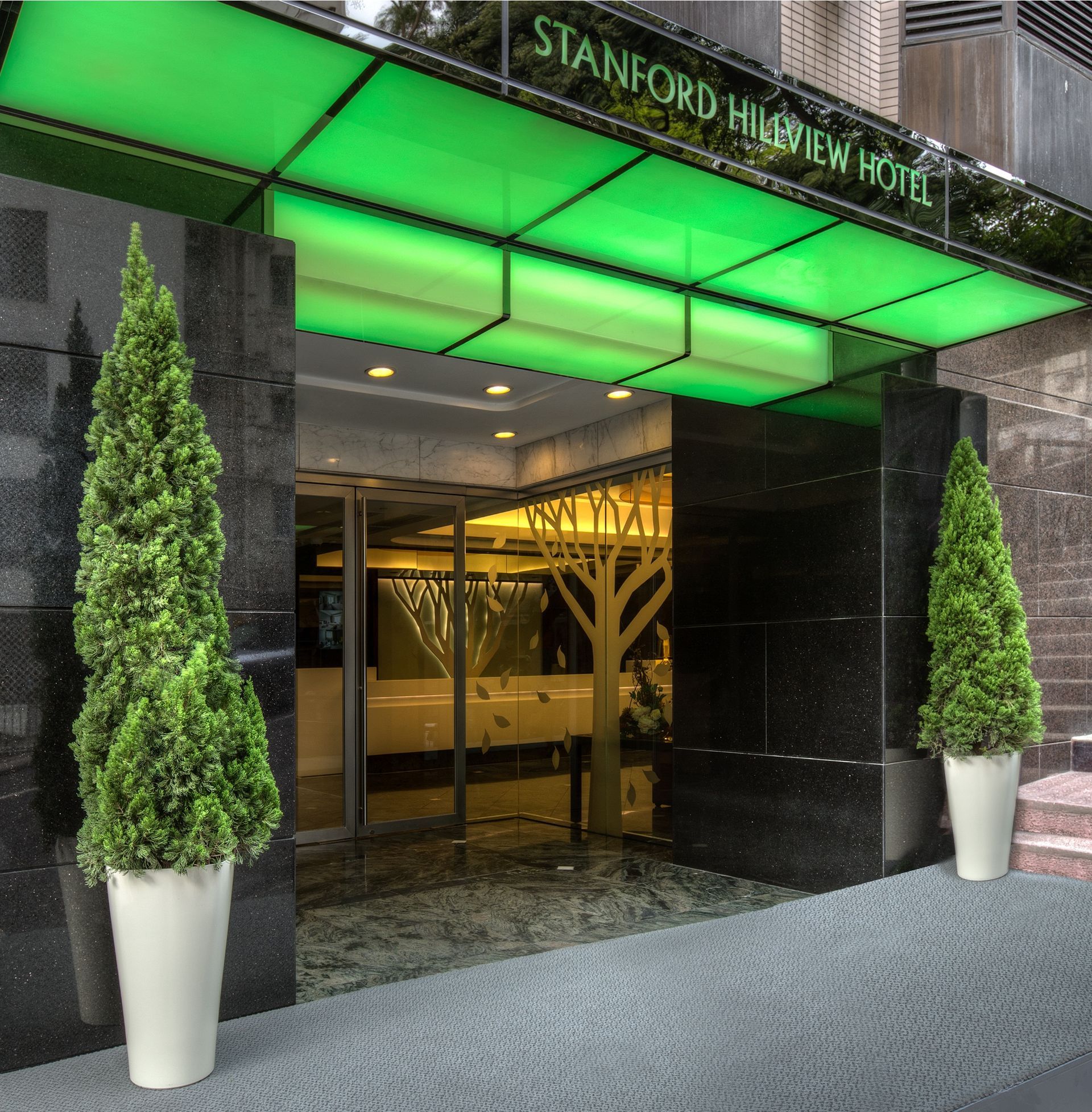 Entrance to the Stanford Hillview Hotel with green canopy, glass doors, potted trees, and stone facade.