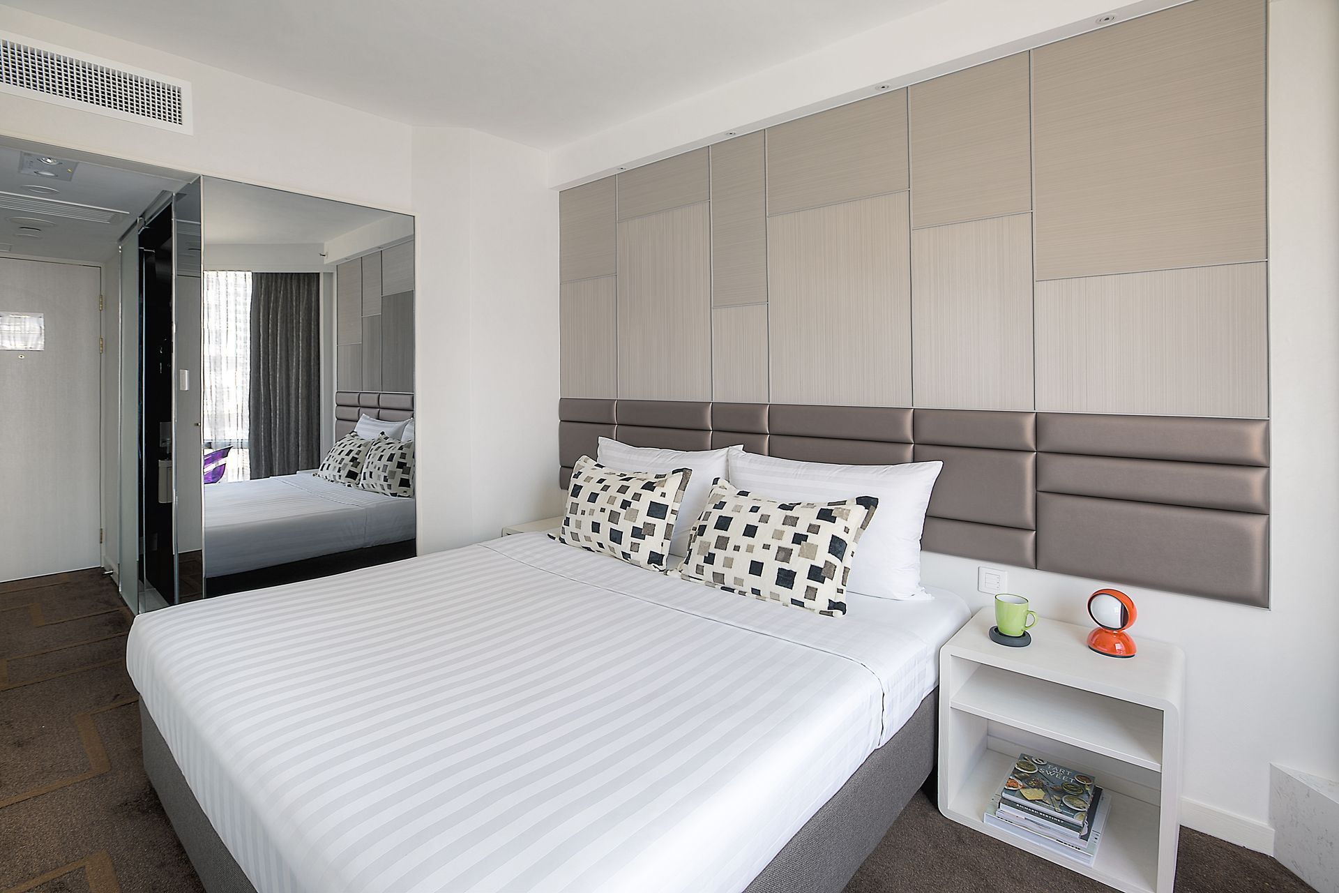 Hotel room with bed, desk, and window overlooking a city. Earth tones dominate.