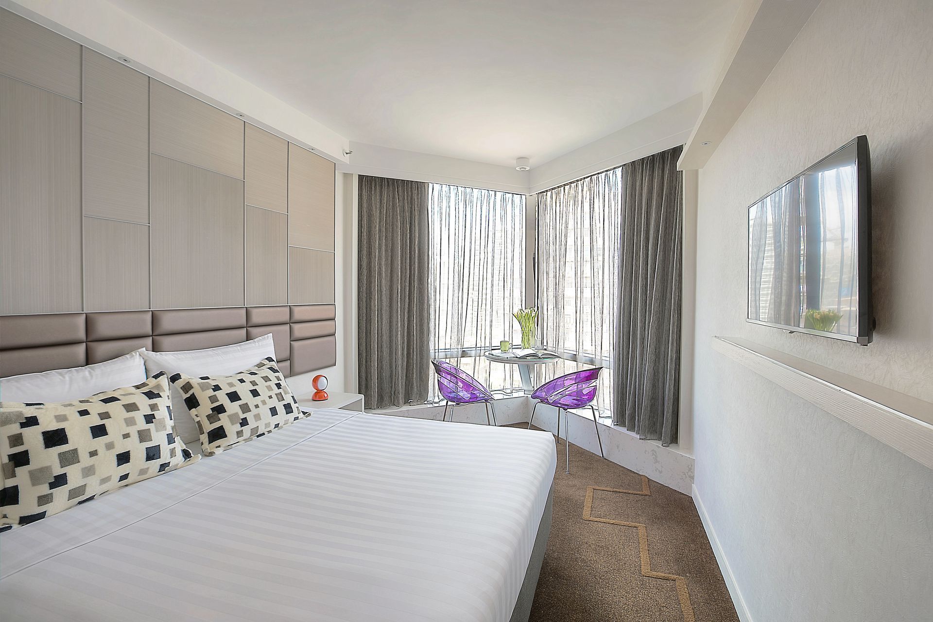 Bedroom with bed, window, and TV. Gray and white decor, two purple chairs.
