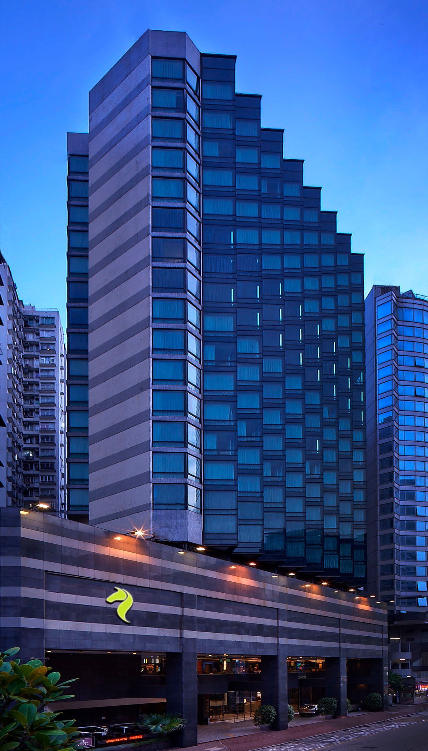 Tall modern building with a multi-tiered facade, glass windows, and a green logo, set against a twilight sky.