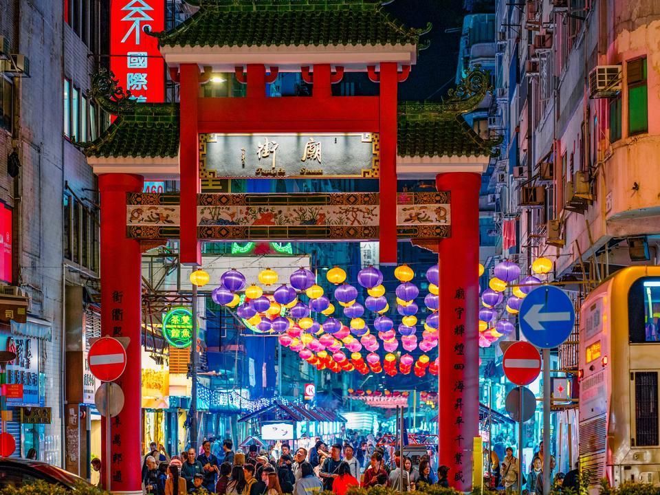Red archway marks the entrance to a bustling Chinatown street at night, adorned with lanterns and neon signs.