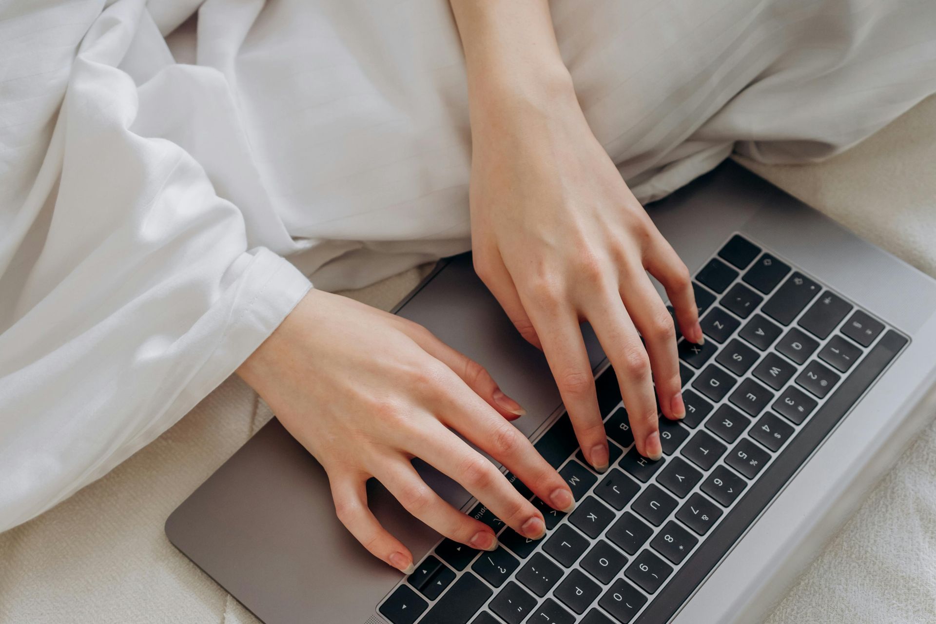 Hands typing on a laptop keyboard, person seated in bed, soft white sheets visible.