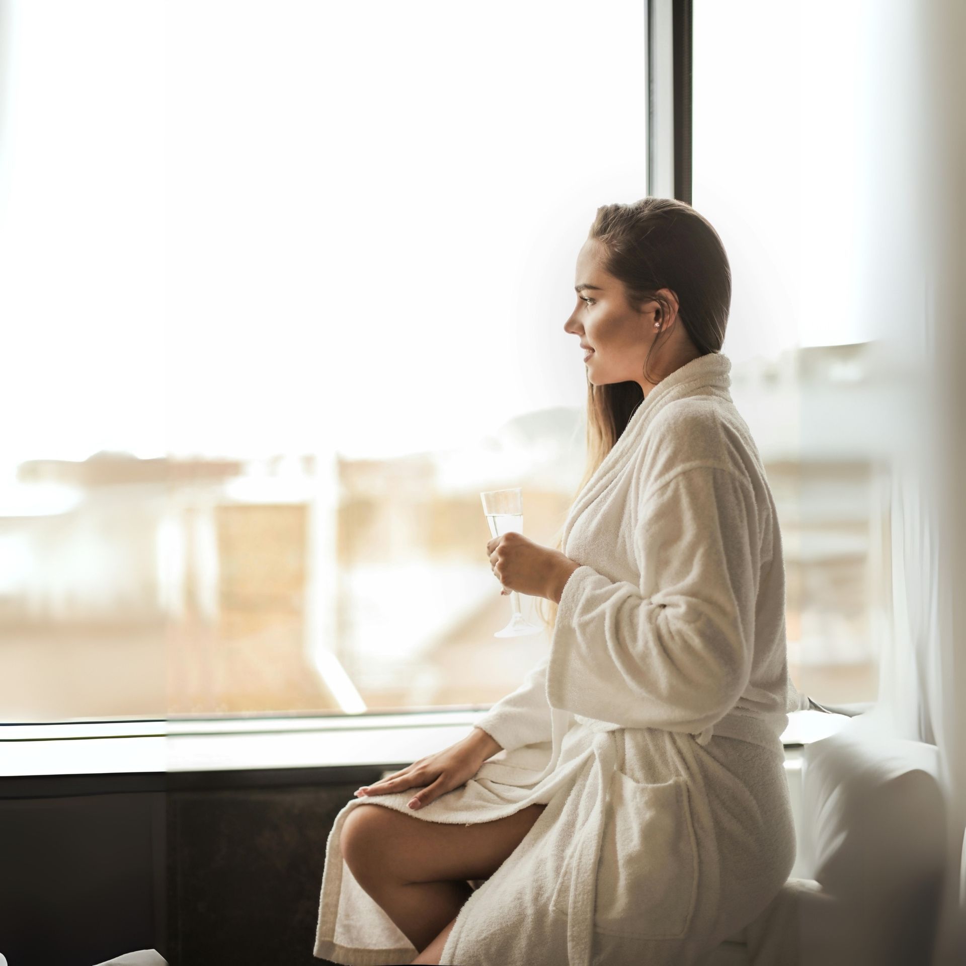 Woman in white robe sits by a window, holding a drink, looking out.