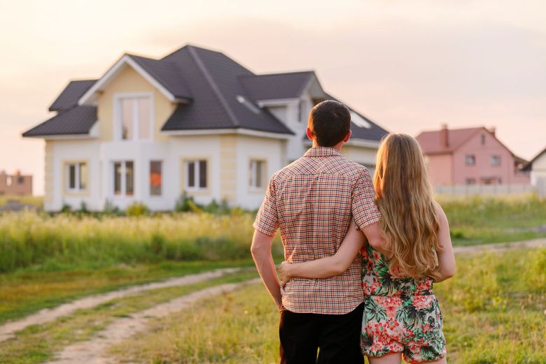 Un couple, bras dessus bras dessous, se tient dans un champ herbeux, regardant vers une maison de banlieue moderne à deux étages au coucher du soleil.