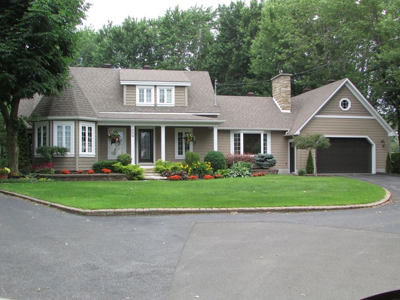 Une maison beige à deux étages avec un toit en bardeaux bruns, un garage sombre et une allée circulaire entourée d'une pelouse verdoyante luxuriante.
