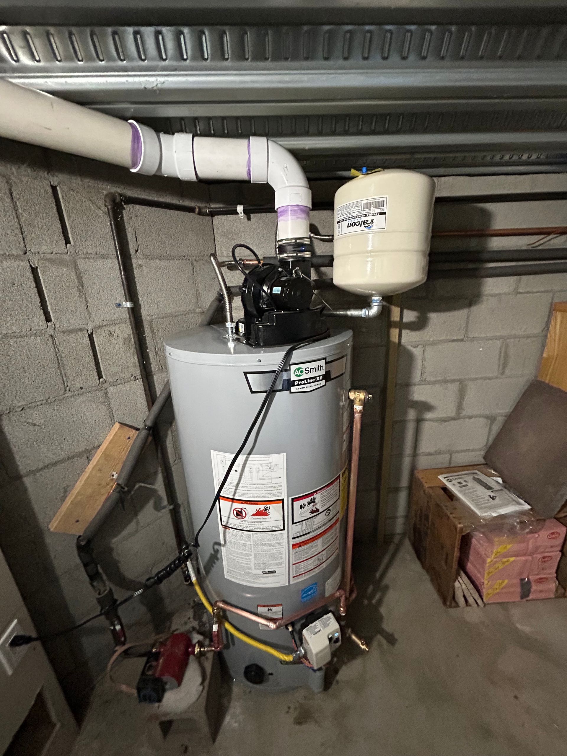 A water heater is sitting in a basement next to a brick wall.