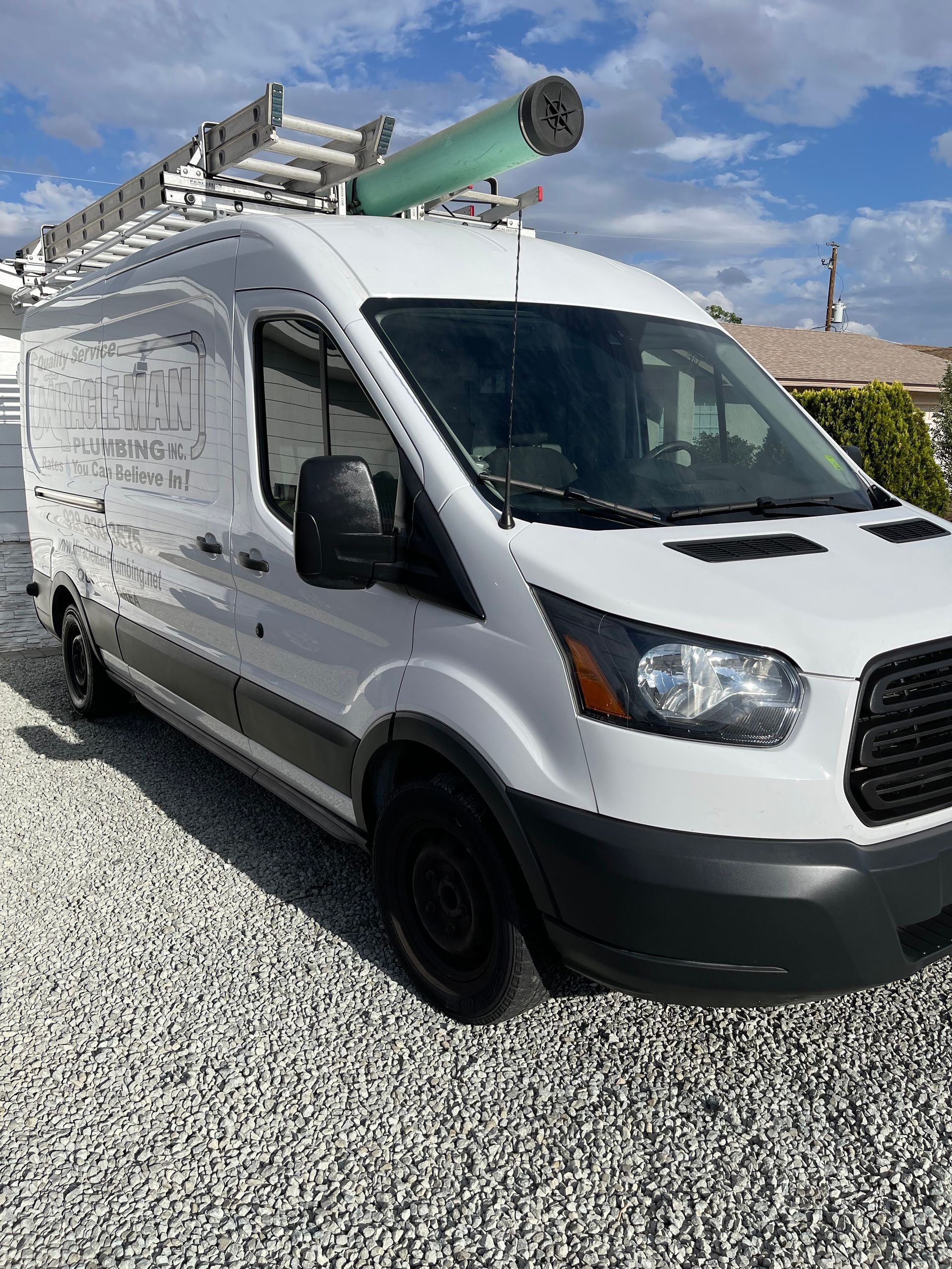 A white van with a ladder on top of it is parked on gravel.
