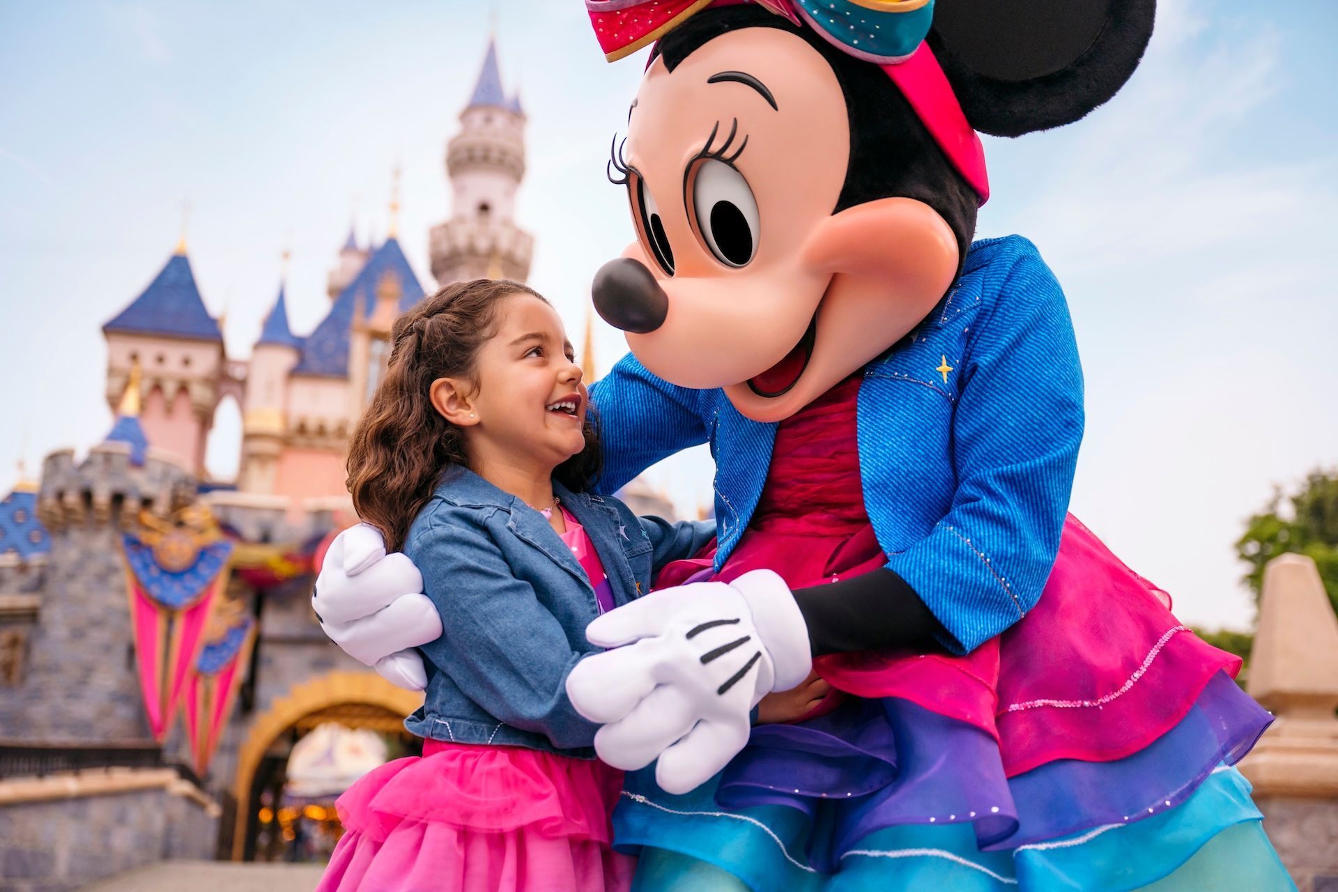 Minnie Mouse hugs a laughing child in front of Sleeping Beauty Castle at Disneyland.
