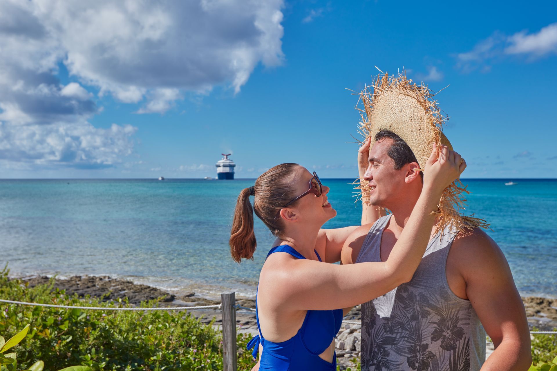 Woman placing a straw hat on a man's head, ocean and cruise ship in background.