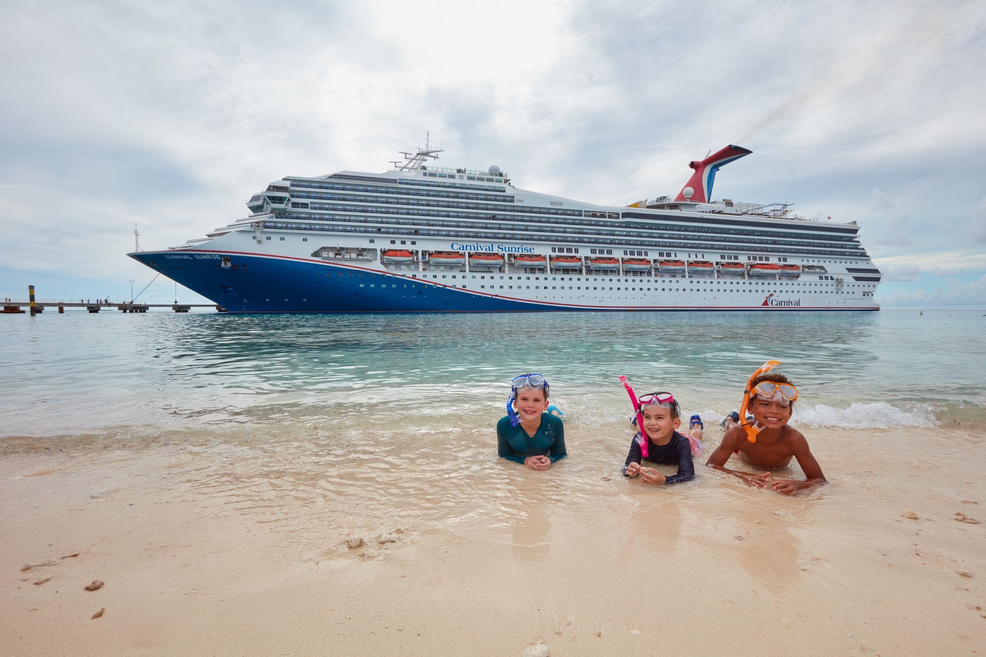 Three children in snorkeling gear on a beach, cruise ship in background.