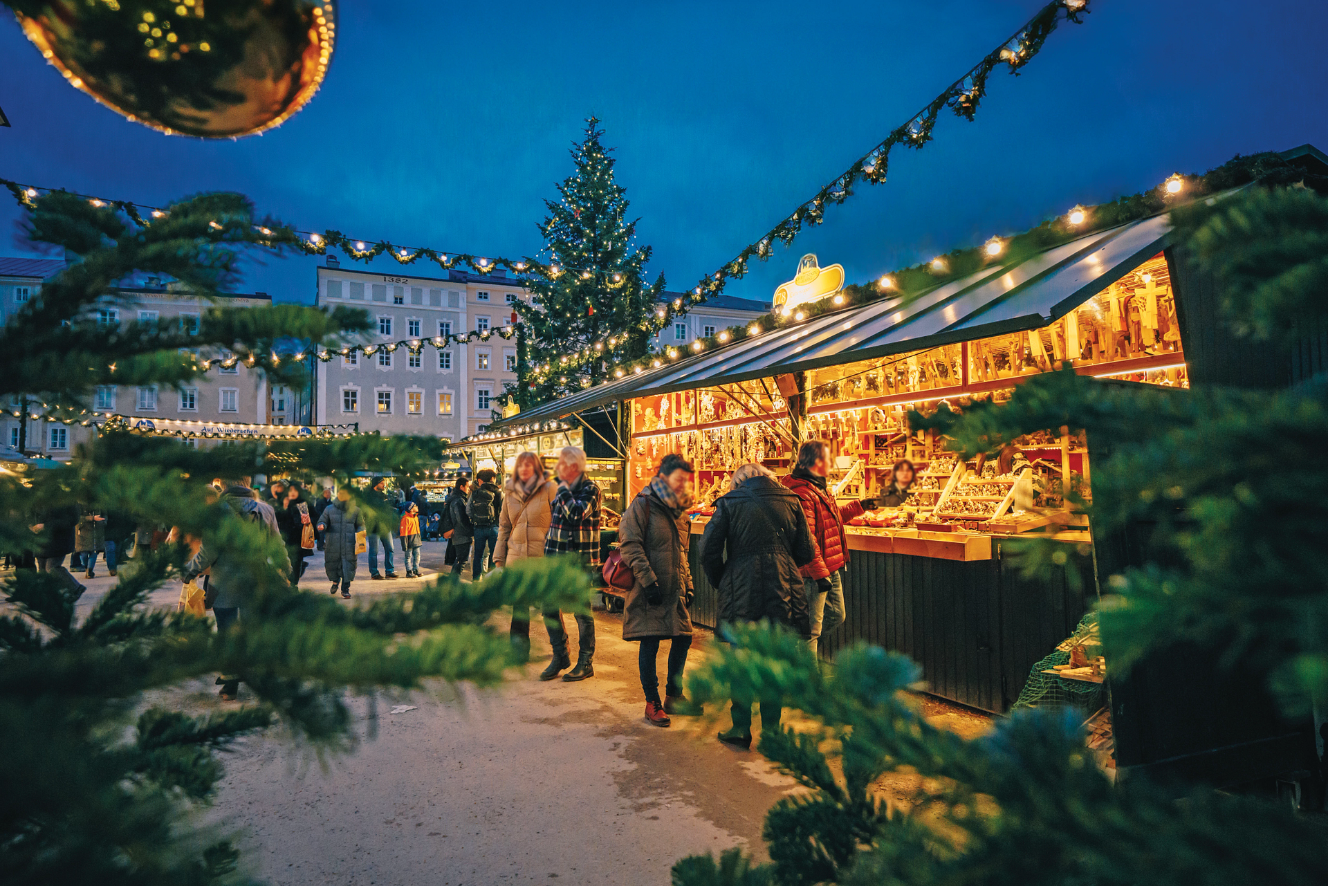 Christmas market at dusk with stalls, people, and a decorated tree.