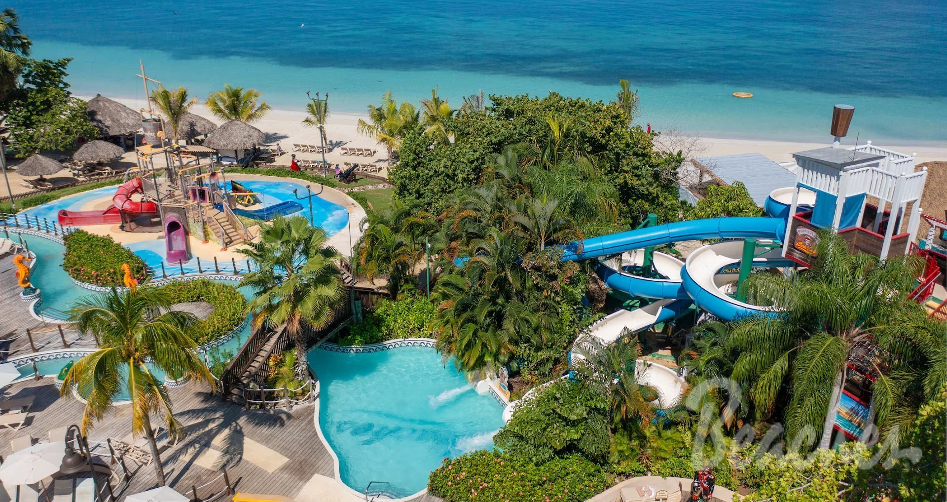 Tropical resort with water slides, pools, beach, and blue ocean. Lush greenery surrounds the water park.