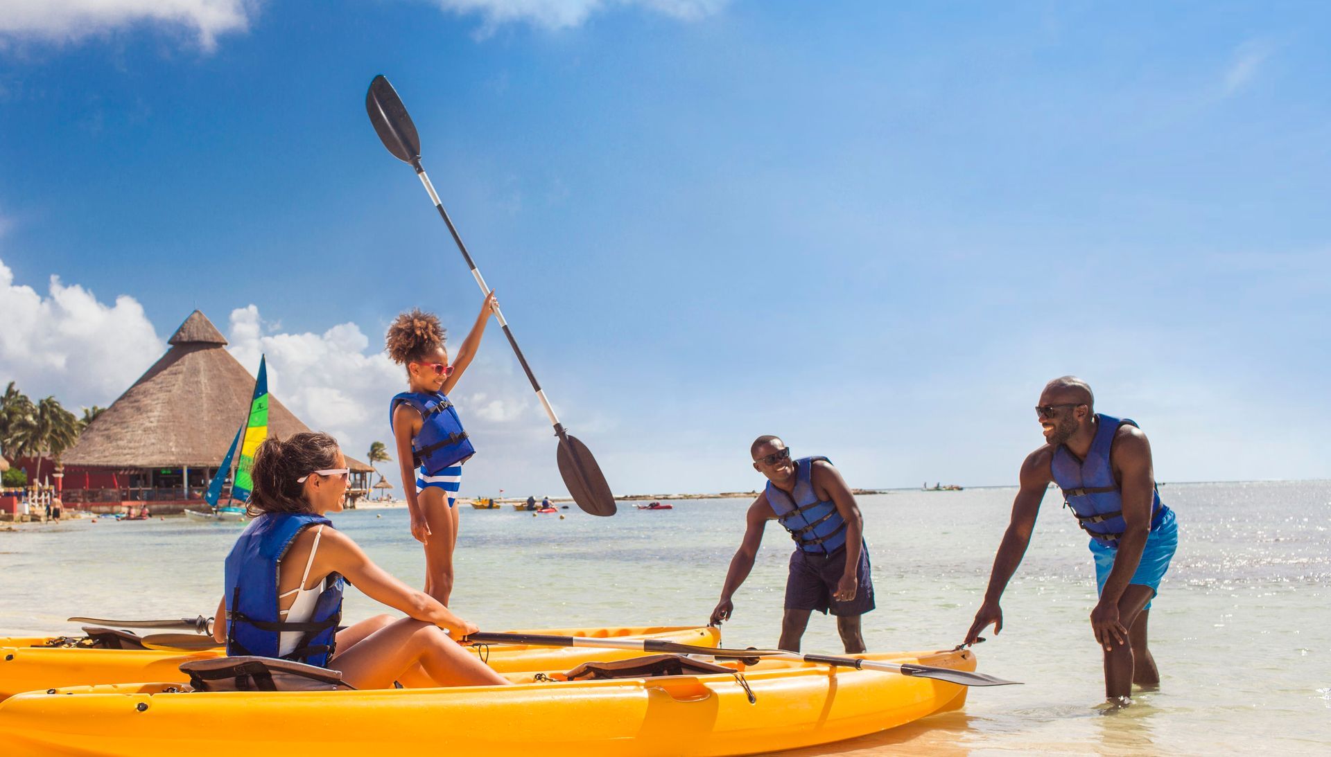 People kayaking on a sunny beach, with a paddler lifting an oar and others wading in shallow water.
