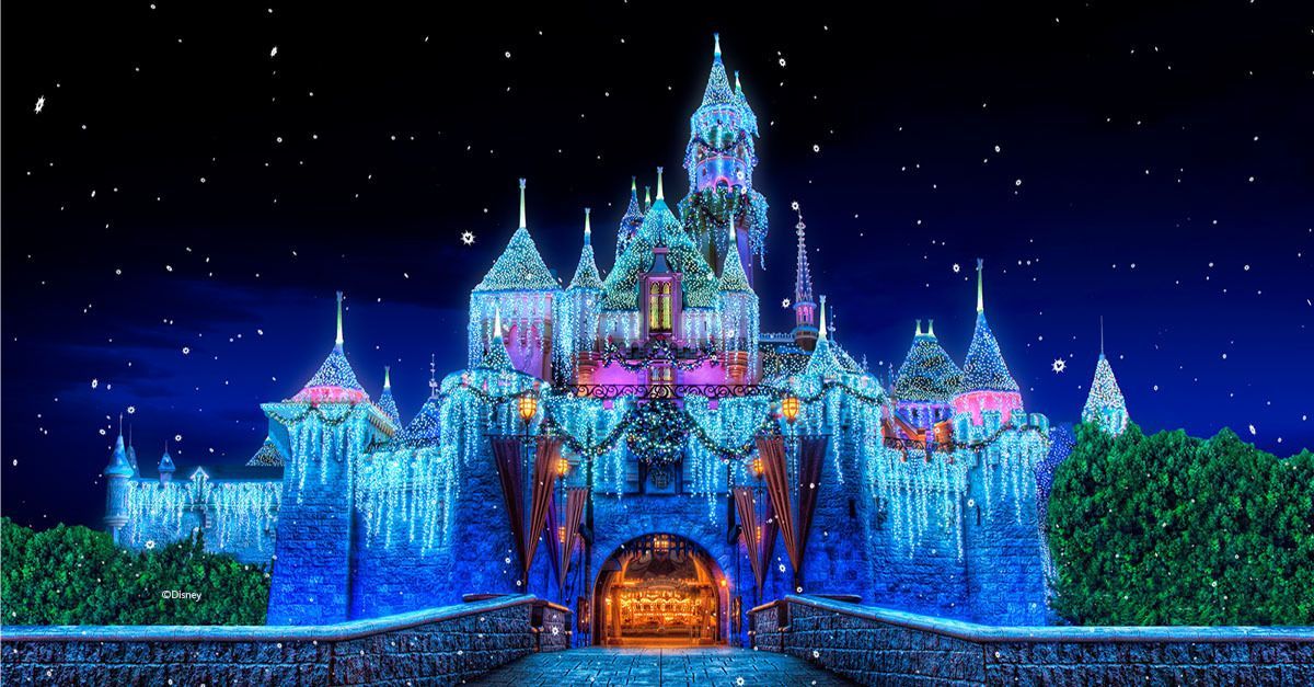 A nighttime view of Sleeping Beauty Castle at Disneyland, illuminated with blue and white holiday lights and snow.