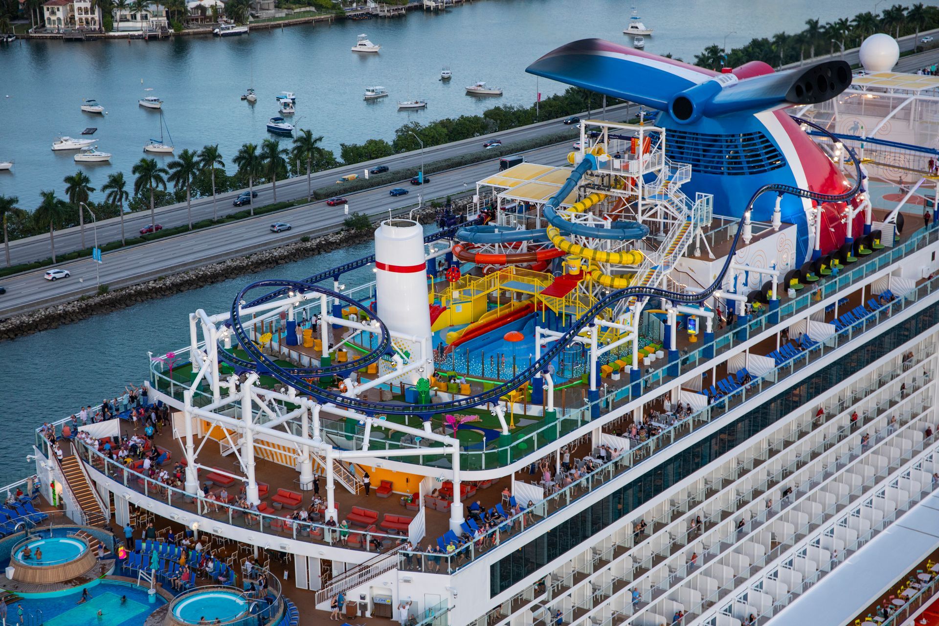 Cruise ship with water slides, roller coaster, and many passengers, docked near a bridge.