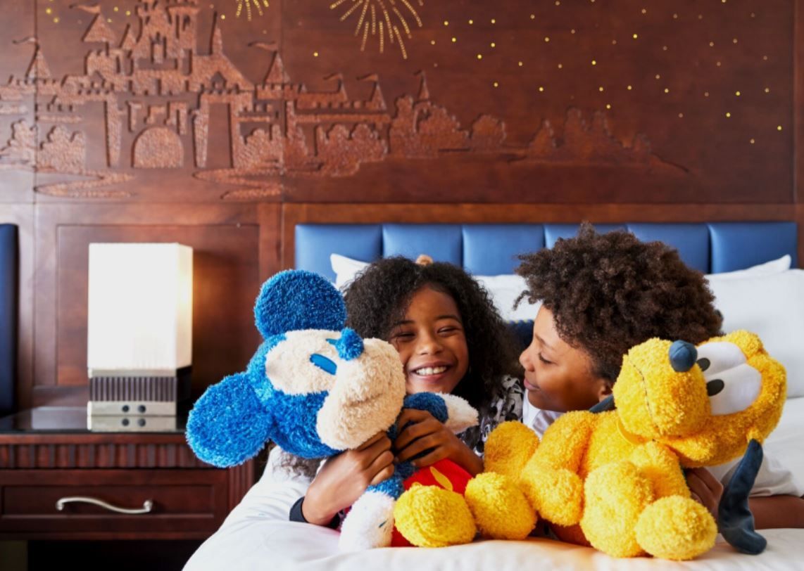 Two children smiling and playing with blue and yellow teddy bears on a bed in a warmly decorated room