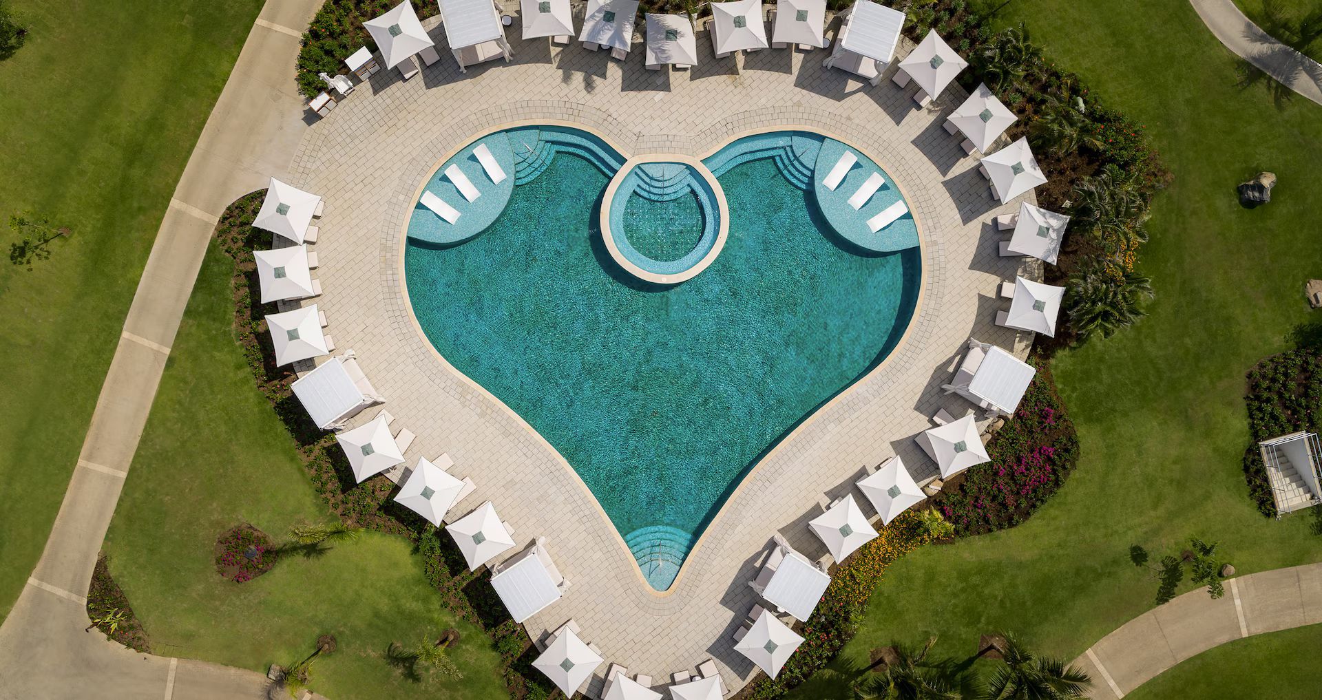 Aerial view of a heart-shaped turquoise swimming pool surrounded by white lounge chairs on a paved patio and green grass.