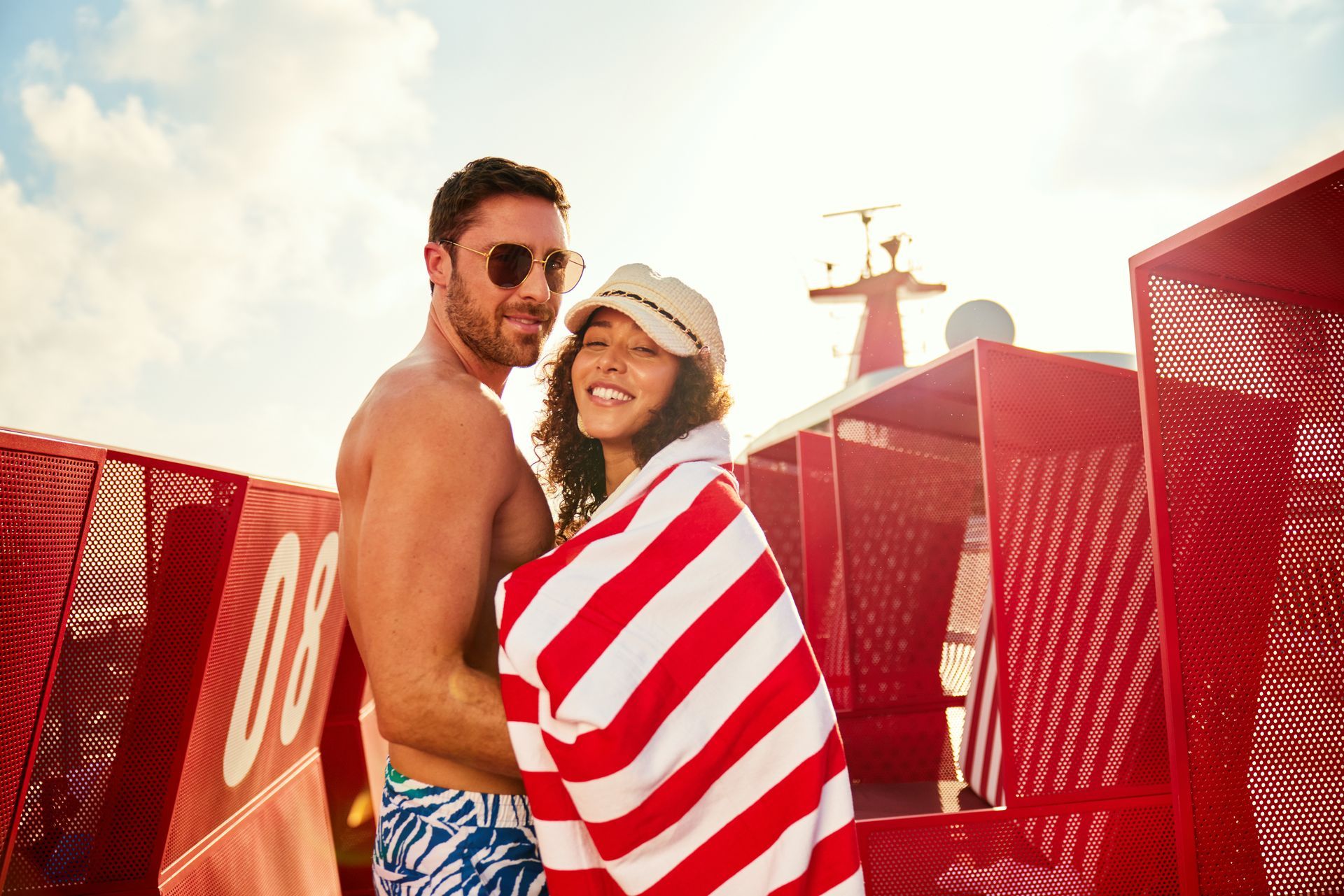 Couple wrapped in striped towel on a deck, leaning against red structures, sunny day.