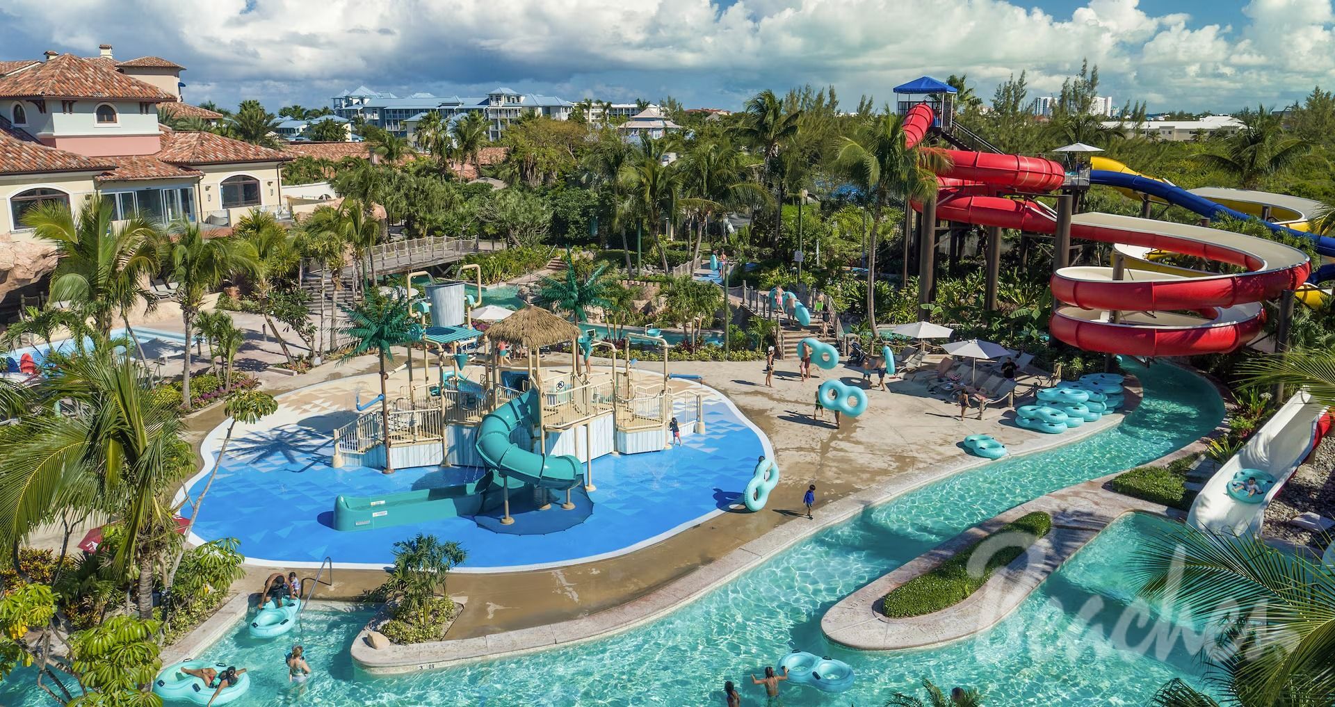 Aerial view of a water park with pools, slides, and lazy river. Blue and red slides, palm trees, and clear water.