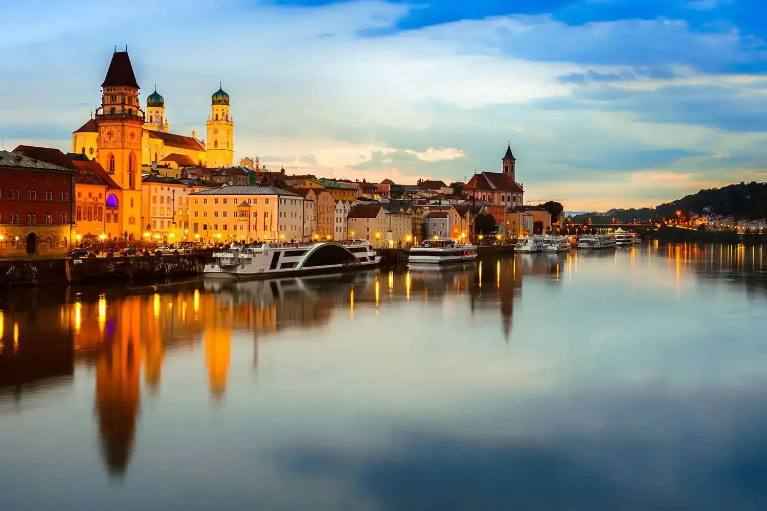 Cityscape of Passau, Germany, at dusk, with illuminated buildings reflected in the calm river.