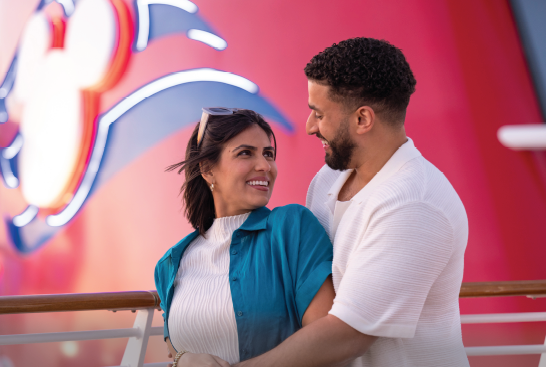 Couple gazing at each other on a cruise ship deck, in front of a red logo.
