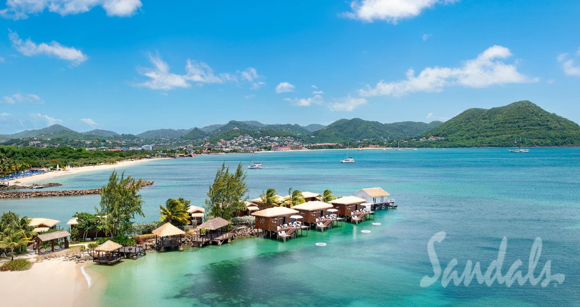 Overwater bungalows on turquoise water, sandy beach, tropical trees, and lush mountains. Sandals resort in St. Lucia.