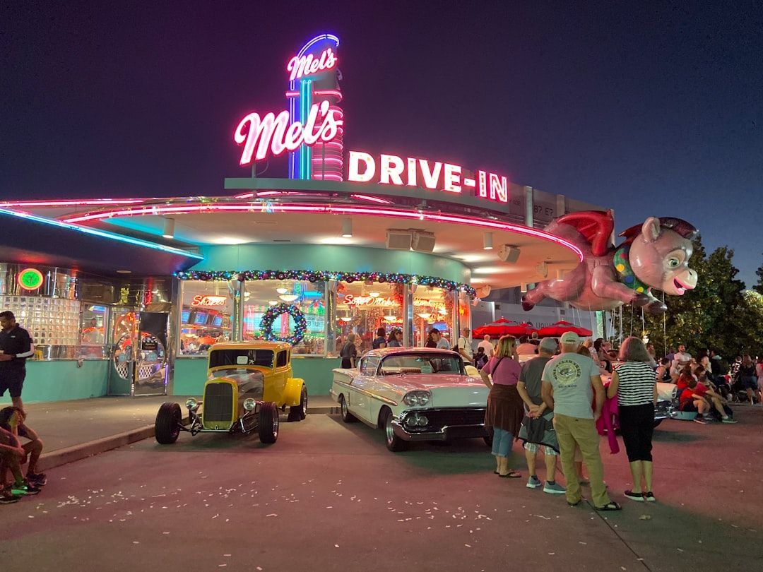 Mel's Drive-In diner at night with classic cars parked in front. Lit sign and decorations, people nearby.