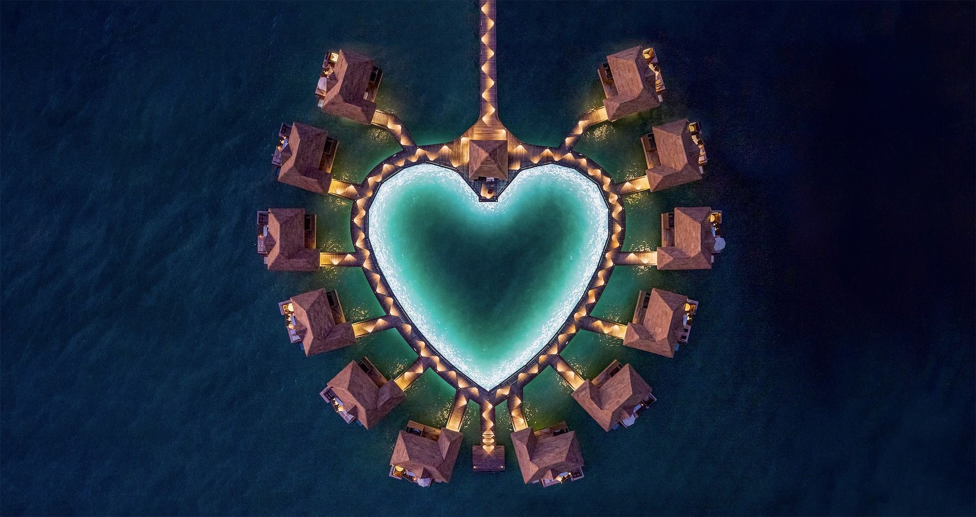 Aerial view of heart-shaped lagoon surrounded by overwater bungalows; illuminated at night.
