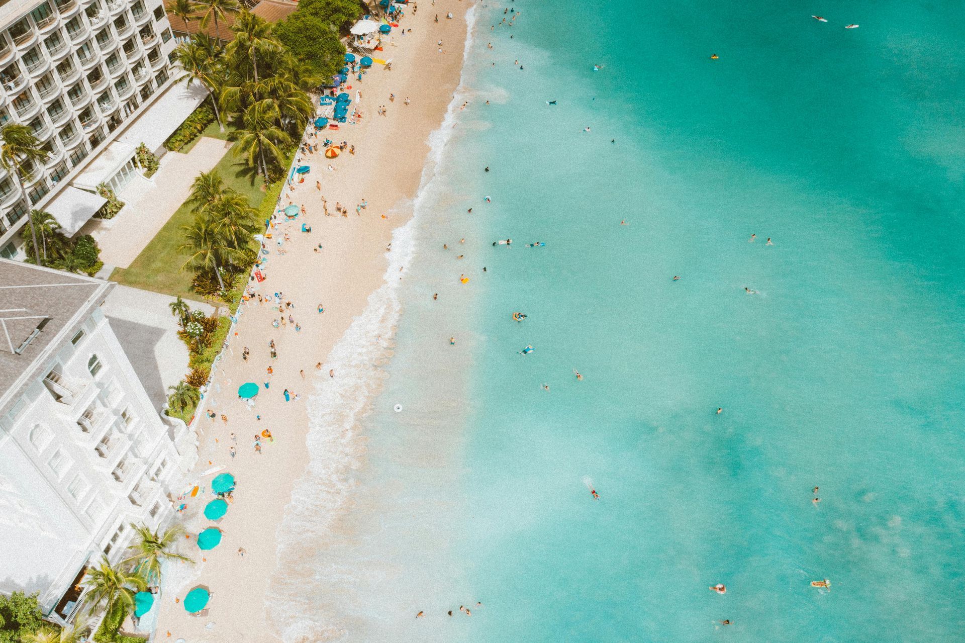 Aerial view of a beach with turquoise water, a sandy shore, and a resort with people swimming and sunbathing.