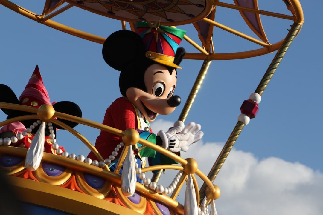 Mickey Mouse in a red jacket and hat waves from a colorful hot air balloon, against a blue sky.