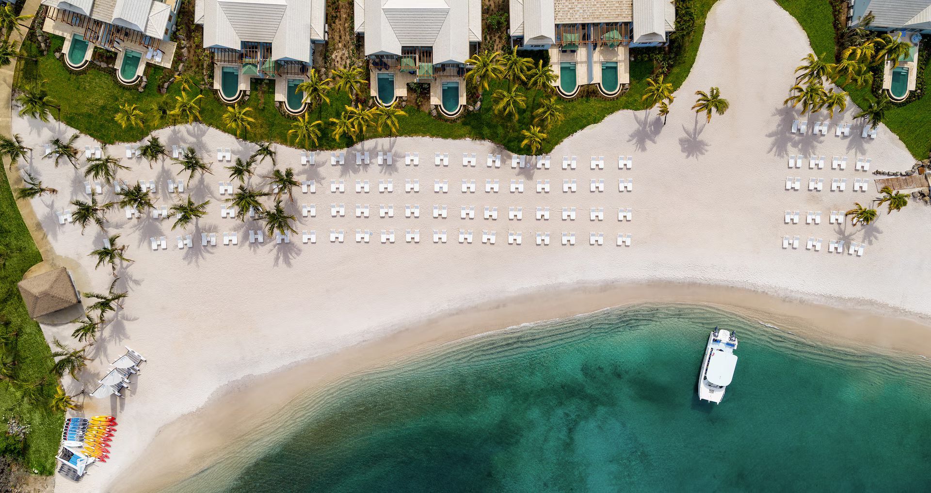 Aerial view of a white sand beach with lounge chairs, turquoise water, and a boat. Lush green vegetation and buildings in the background.
