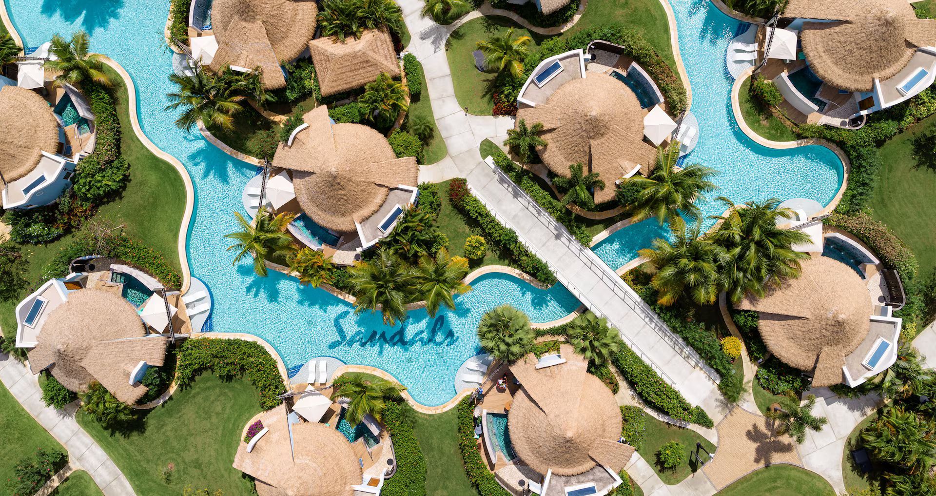 Aerial view of a tropical resort with round thatched-roof huts and a flowing blue pool.