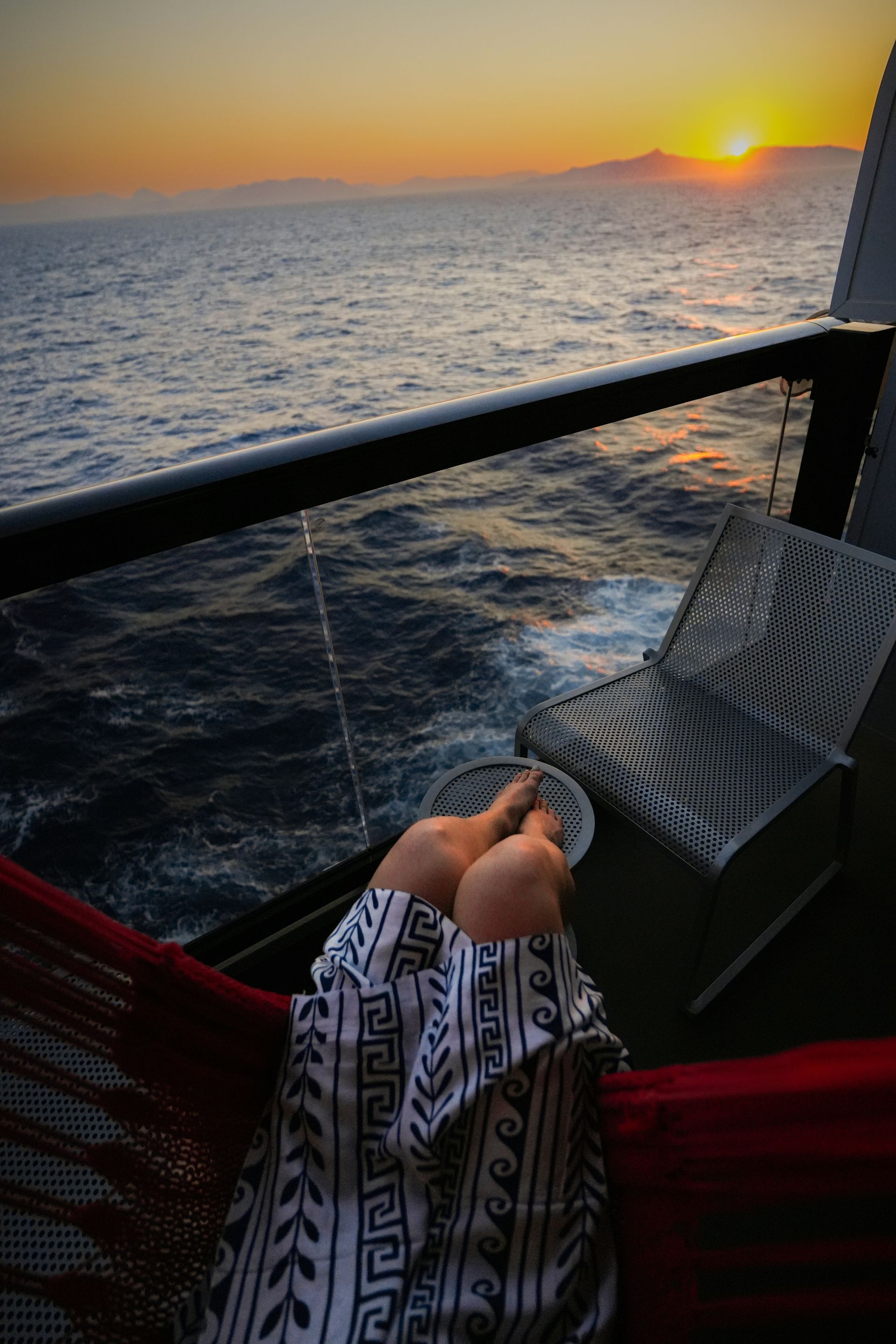 Person relaxing in hammock on a balcony, watching sunset over ocean.