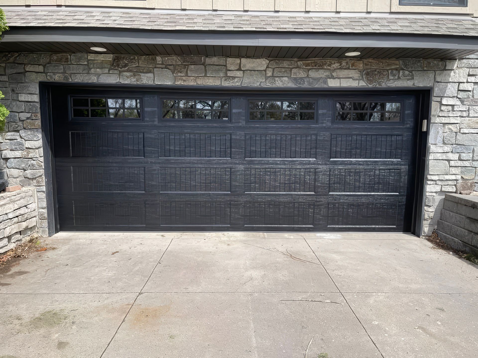 A black, four-section garage door with small windows at the top, set in a stone facade with a concrete driveway.
