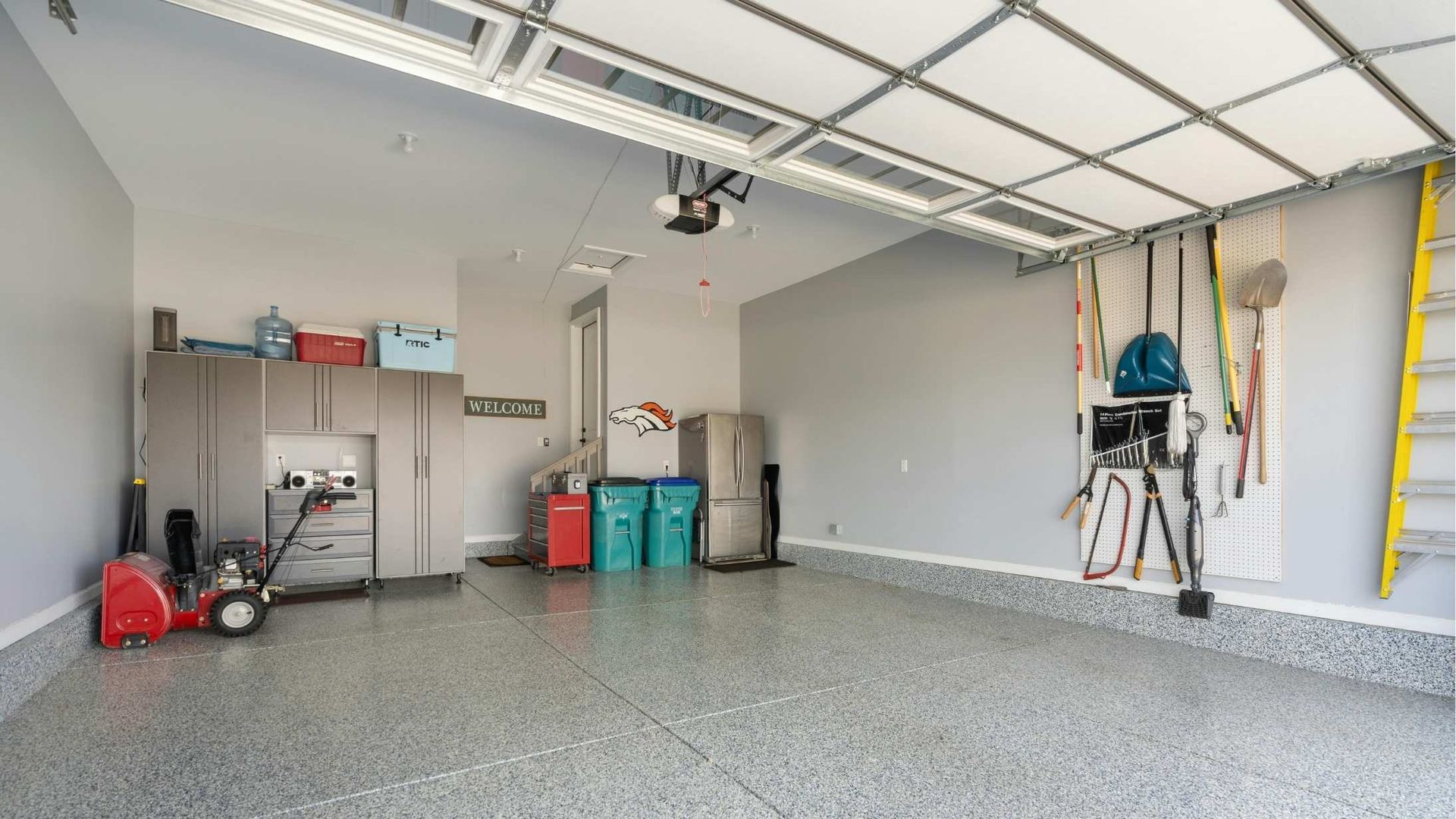 An organized two-car garage with gray walls, speckled epoxy floors, gray storage cabinets, and a yellow ladder.