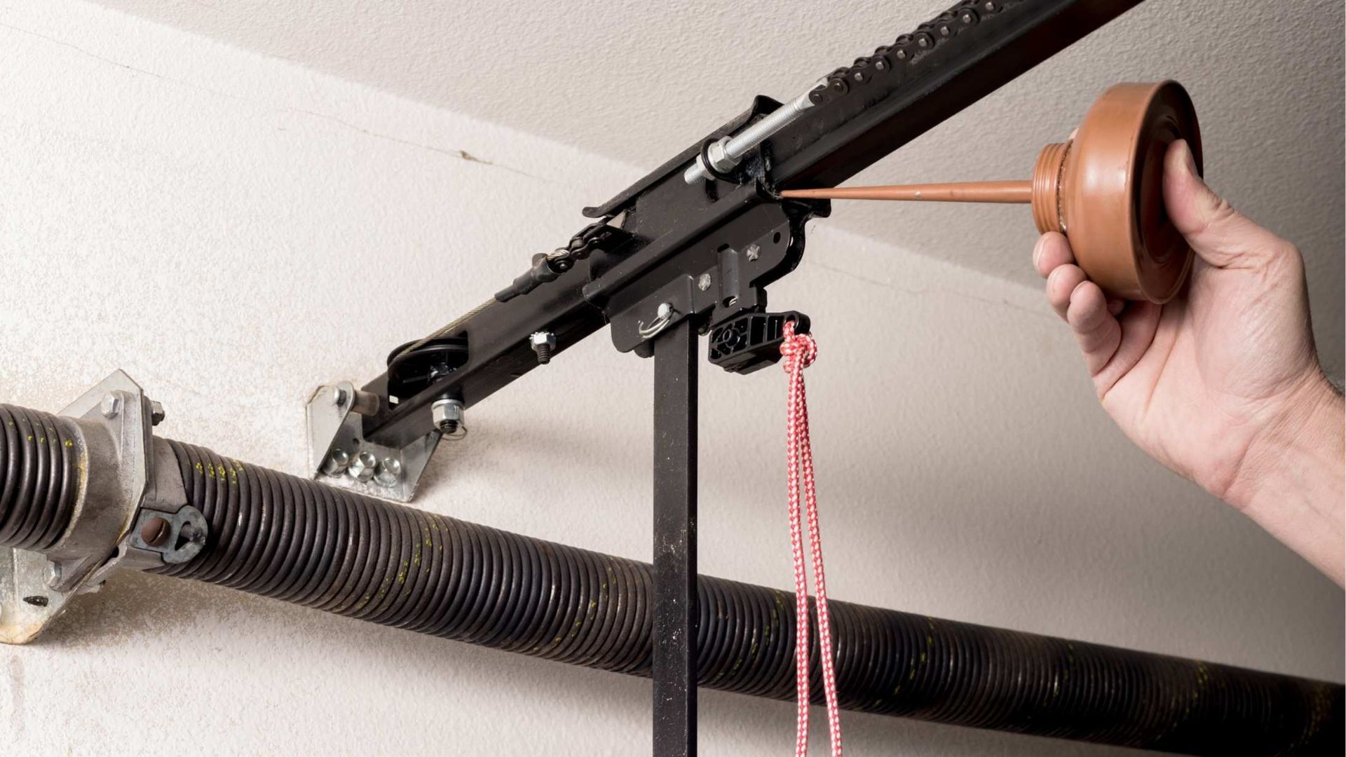 A hand uses an oil can to lubricate a garage door opener rail and trolley system.