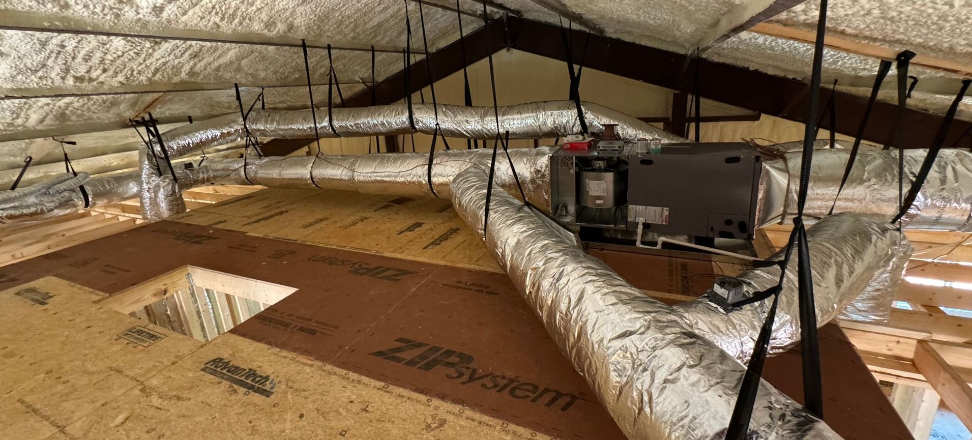 HVAC ductwork installation with foam-insulated ceiling in attic space, showing flexible air ducts and mounted air handler during system upgrade.