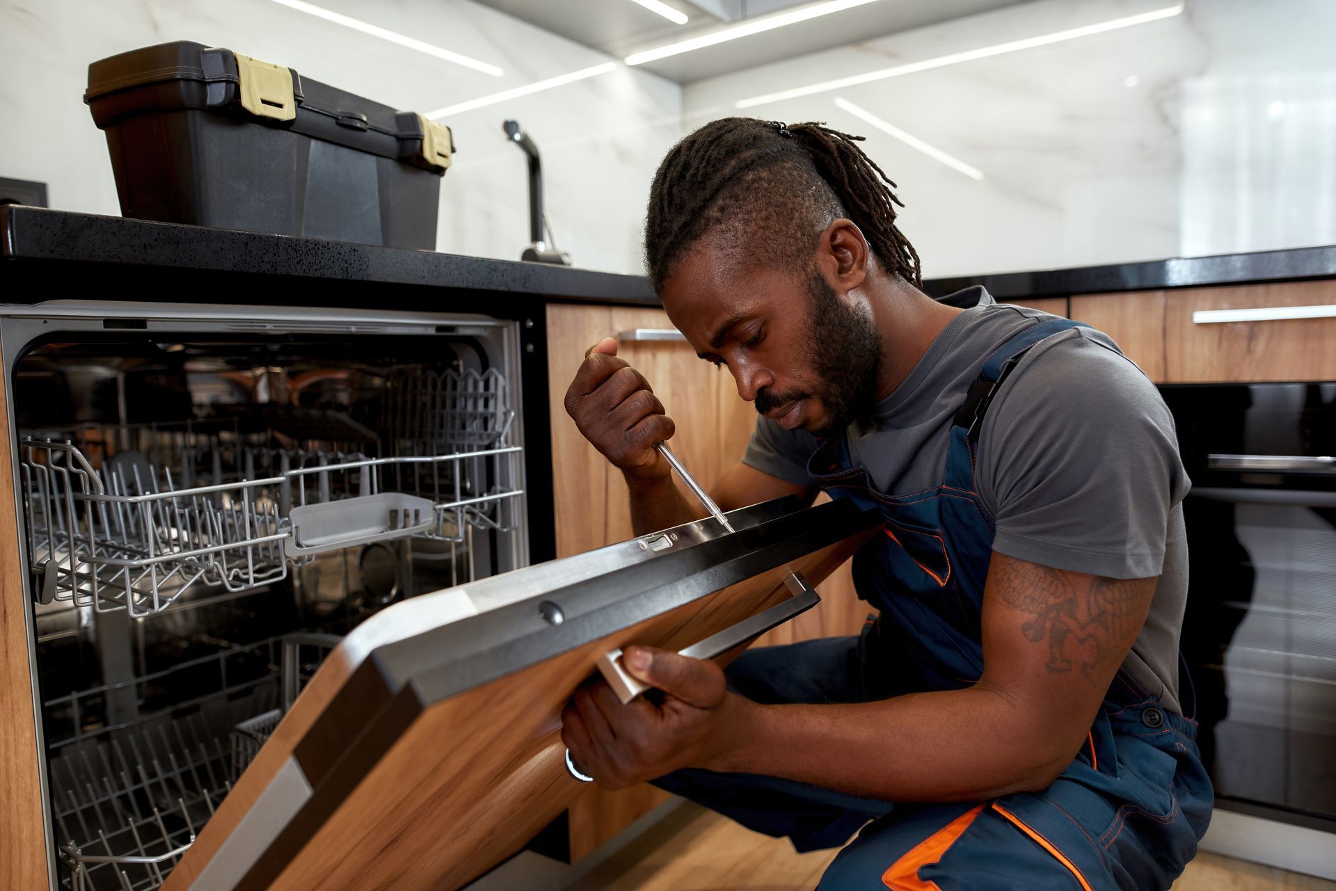 A technician in work coveralls uses a screwdriver to repair the open door of a dishwasher in a kitchen.