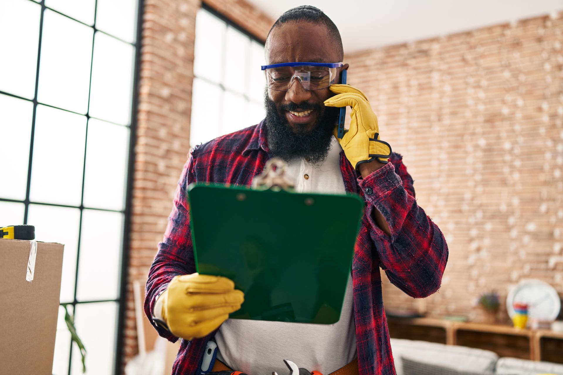 A person wearing safety goggles and work gloves looks at a clipboard while talking on a smartphone in a room.