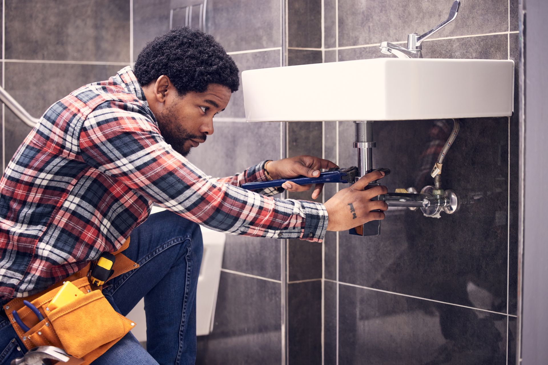 A person wearing a plaid shirt and tool belt works on the plumbing beneath a wall-mounted sink in a tiled bathroom.